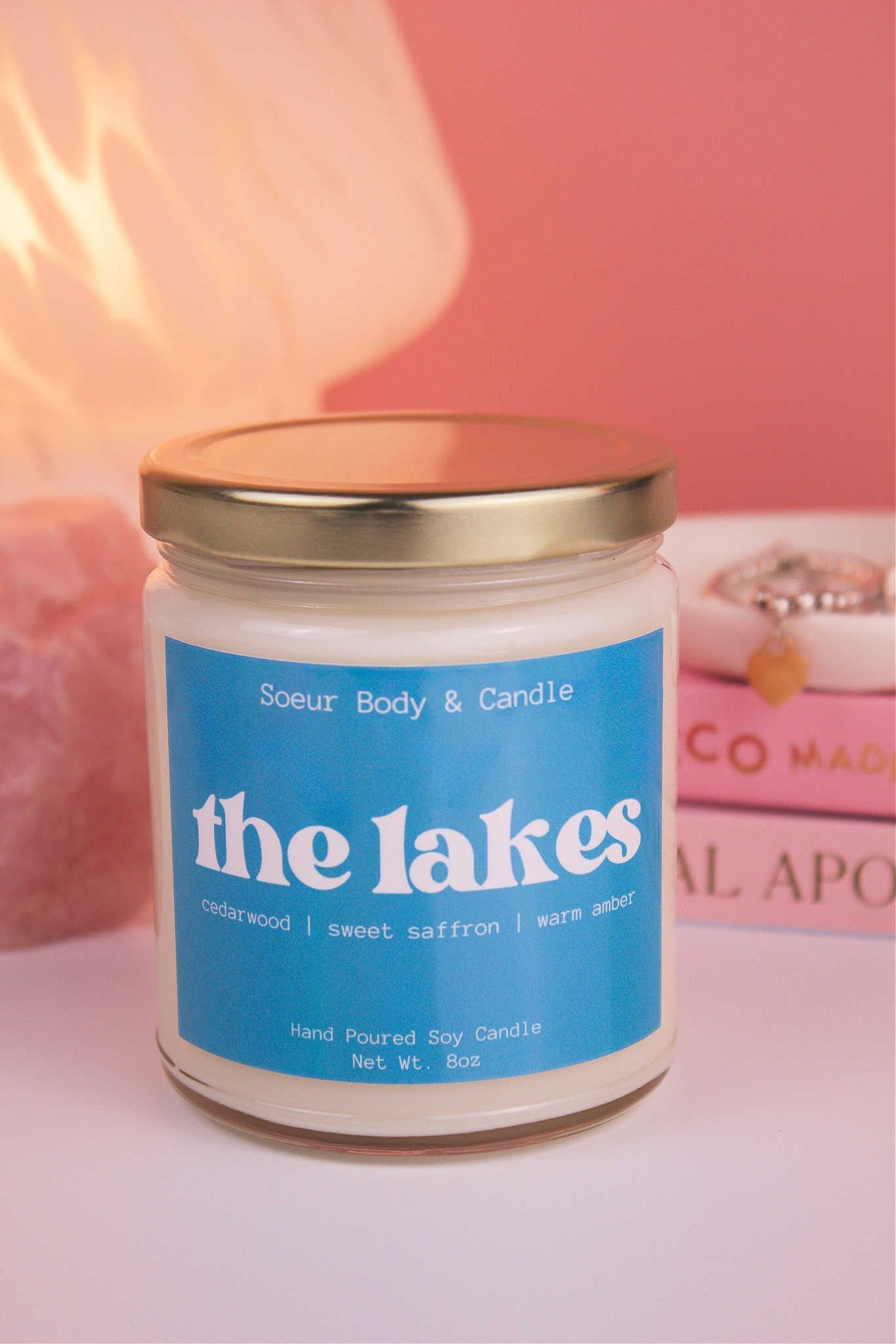 Close-up of The Lakes candle with the lid on, with a stack of books and a jewelry dish in the background, set against a lamp and crystals; fragrance notes: cedarwood, sweet saffron, and warm amber.