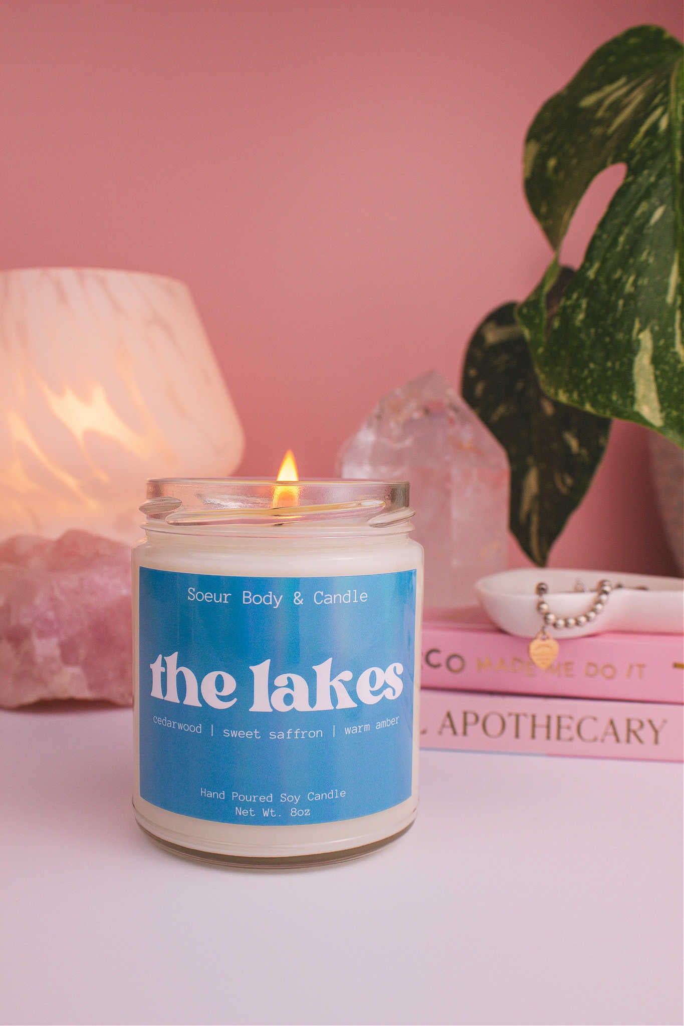 The Lakes candle lit, with a stack of books and a jewelry dish in the background, set against a lamp, plant, and crystals; fragrance notes: cedarwood, sweet saffron, and warm amber.