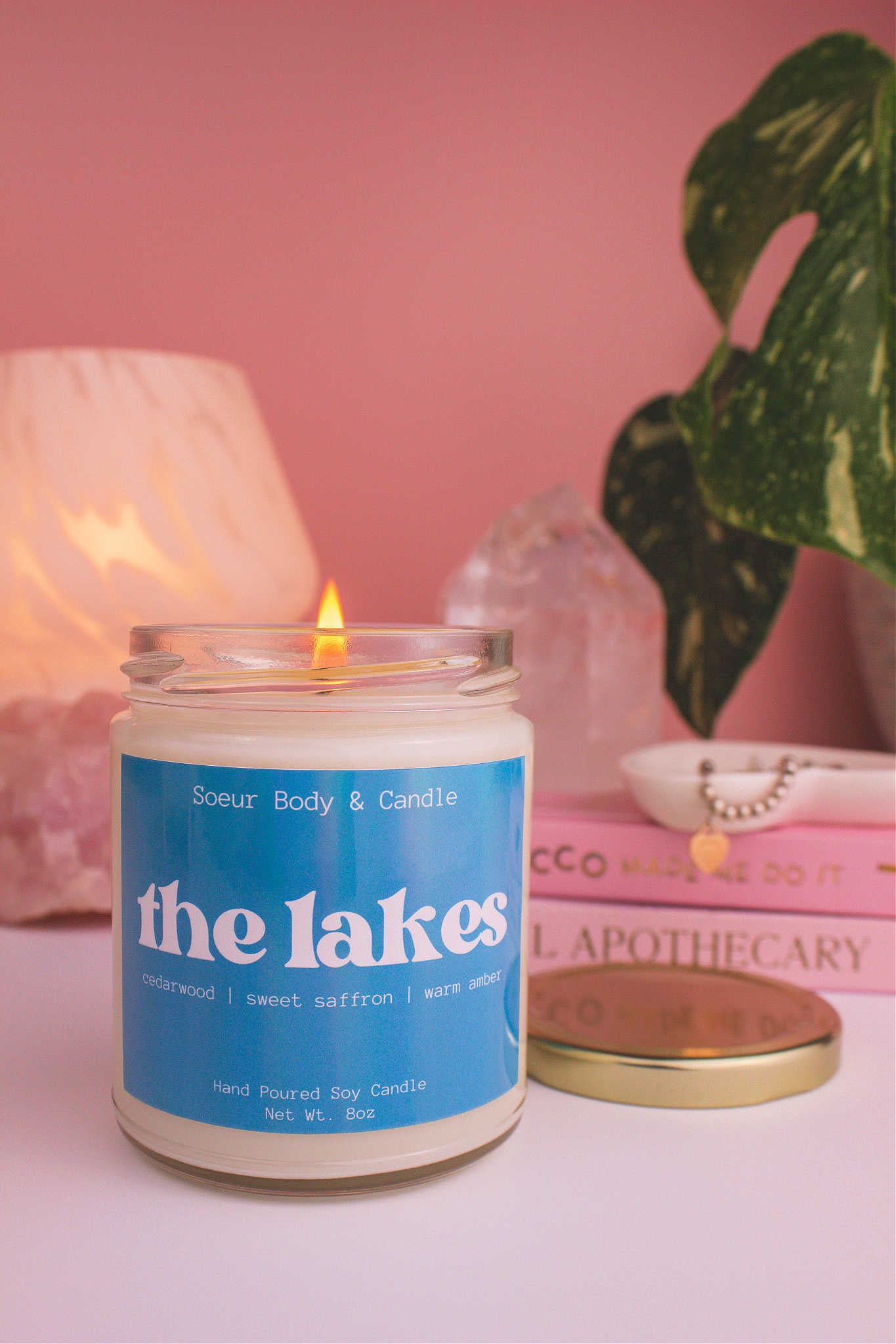 Close-up of The Lakes candle lit, with a stack of books and a jewelry dish in the background, set against a lamp, plant, and crystals; fragrance notes: cedarwood, sweet saffron, and warm amber.