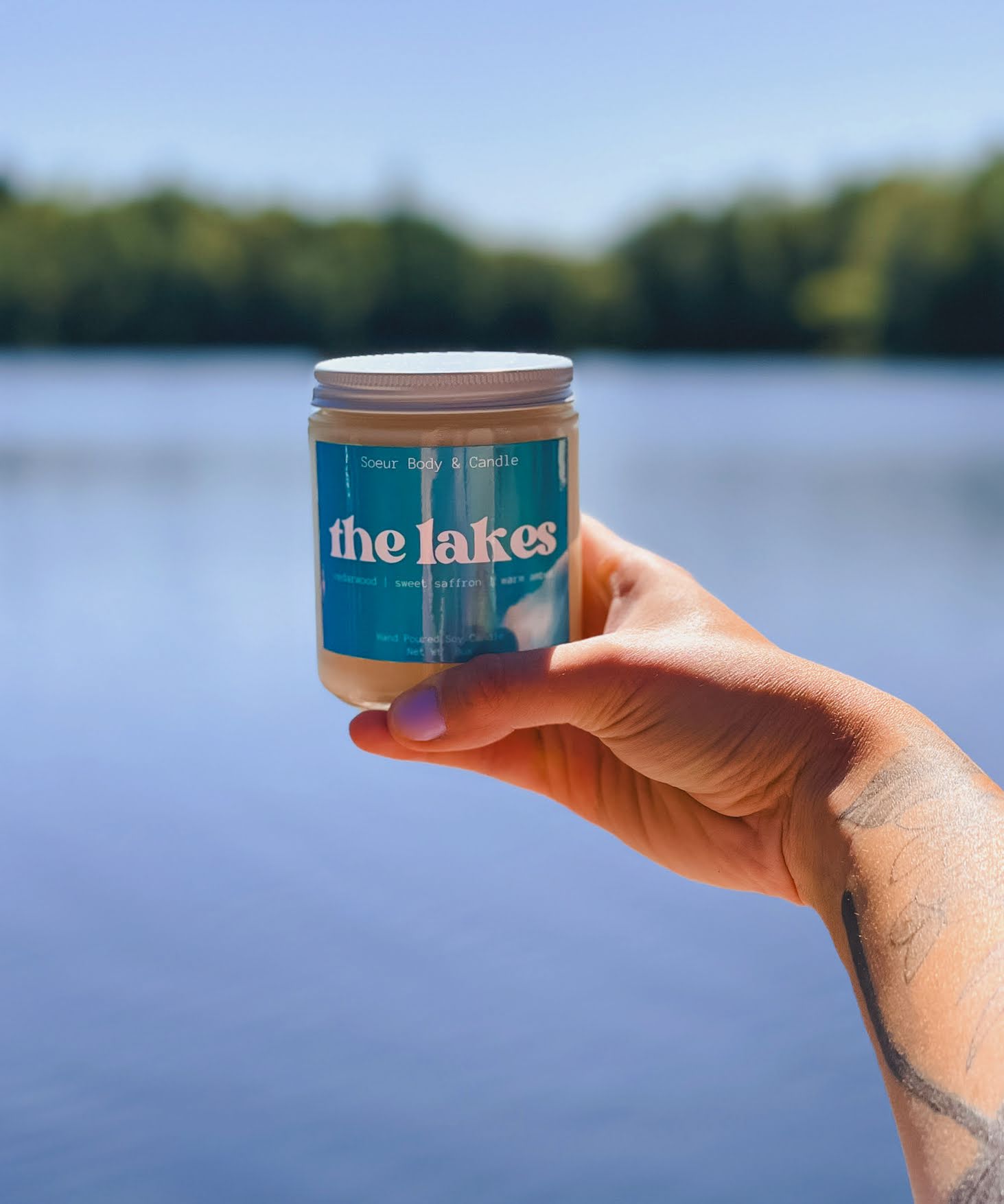 The Lakes Candle held in hand, against a blurred background of a lake and tress in the distance. Fragrance notes: cedarwood, sweet saffron, and warm amber.
