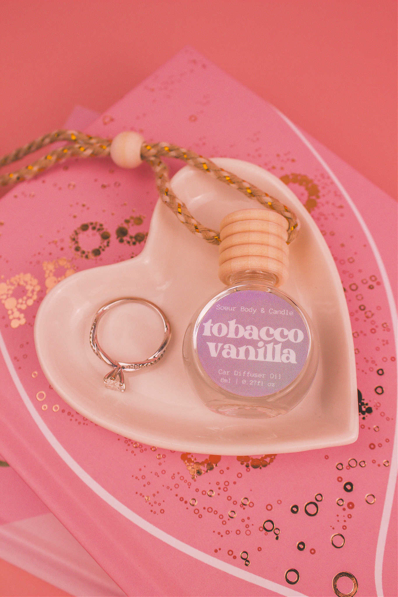 Tobacco Vanilla car diffuser in a jewelry dish with a ring beside it, sitting on top of books, featuring notes of vanilla, amber, tobacco leaves, and tonka bean.