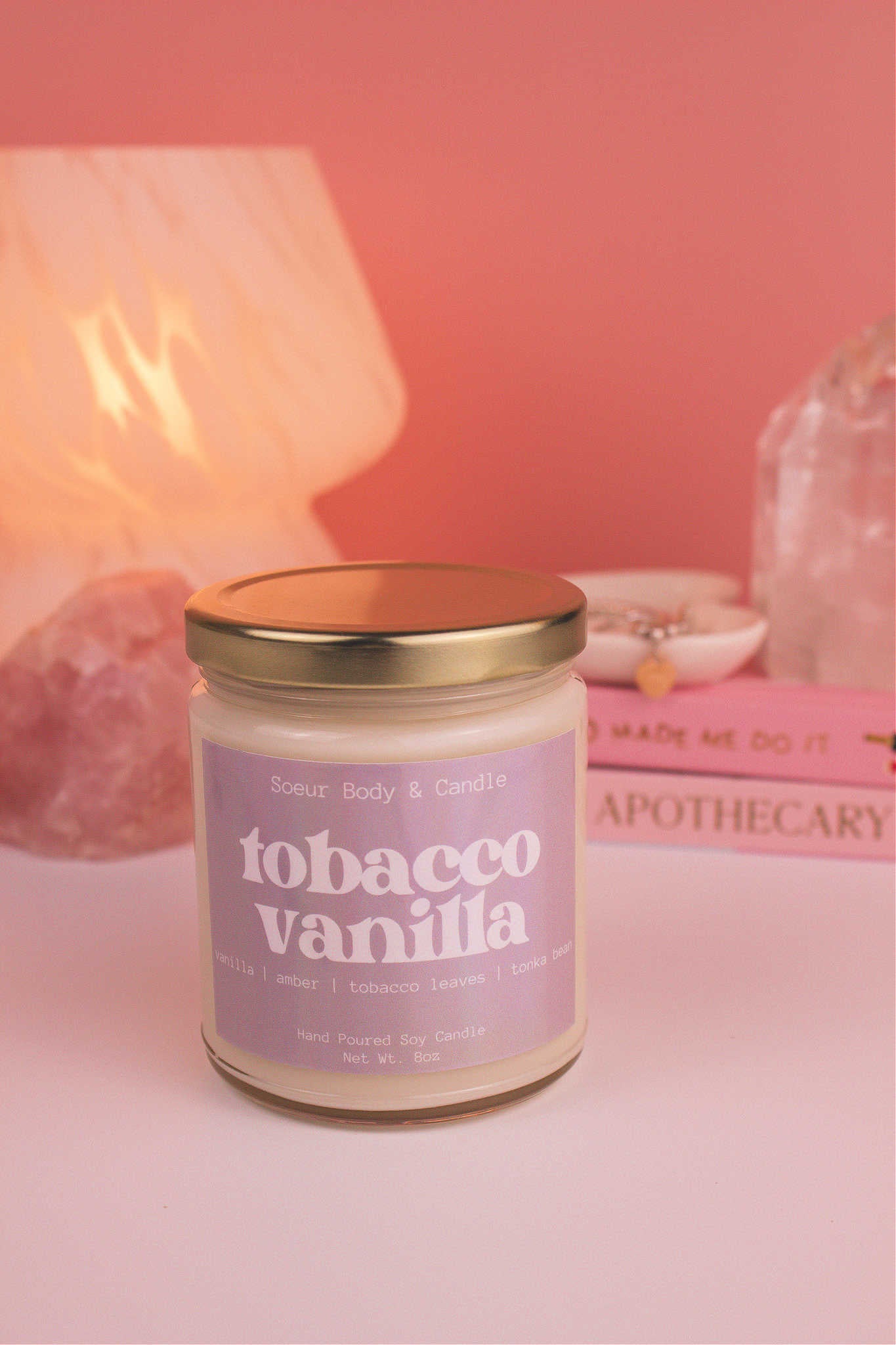 Tobacco Vanilla candle with the lid on, with a stack of books and a jewelry dish in the background, set against a lamp and crystals; fragrance notes: vanilla, amber, tobacco leaves, and tonka bean.