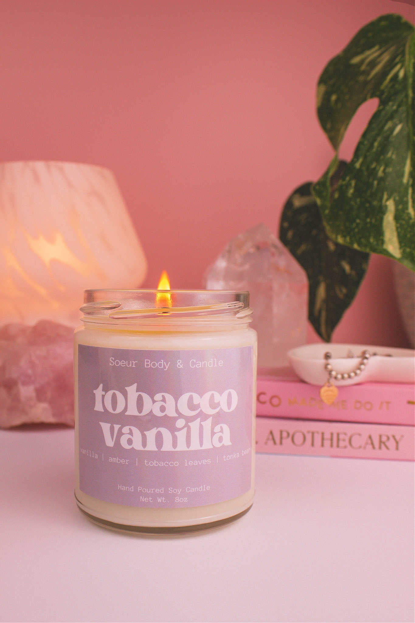 Tobacco Vanilla candle lit, with a stack of books and a jewelry dish in the background, set against a lamp, plant, and crystals; fragrance notes: vanilla, amber, tobacco leaves, and tonka bean.