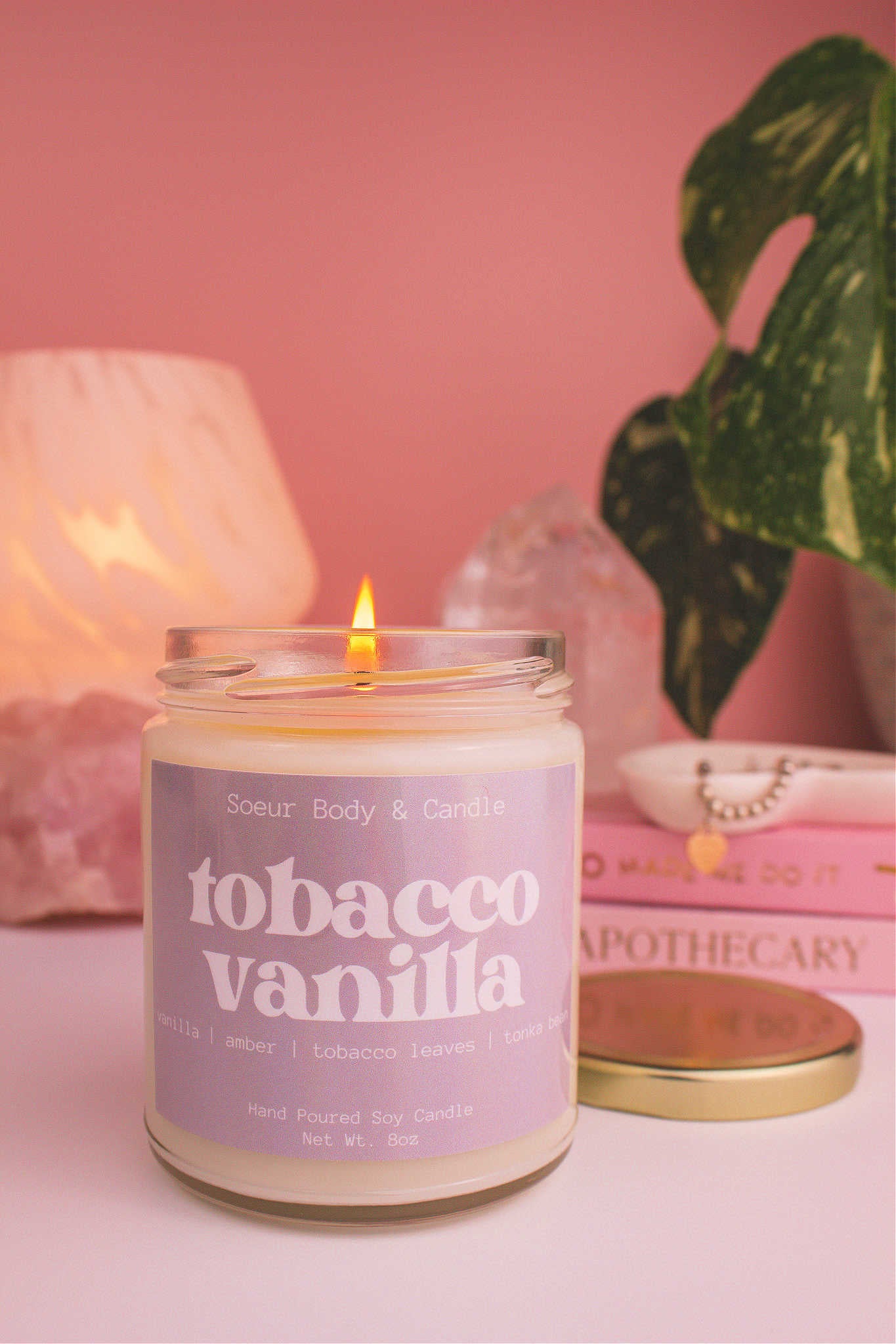Close-up of Tobacco Vanilla candle lit, with a stack of books and a jewelry dish in the background, set against a lamp, plant, and crystals; fragrance notes: vanilla, amber, tobacco leaves, and tonka bean.