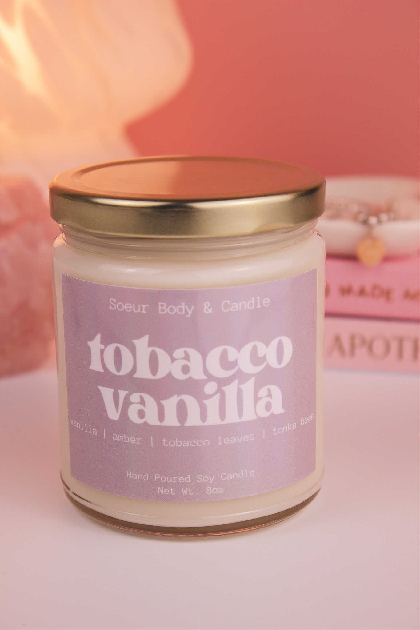 Close-up of Tobacco Vanilla candle with the lid on, with a stack of books and a jewelry dish in the background, set against a lamp and crystals; fragrance notes: vanilla, amber, tobacco leaves, and tonka bean.