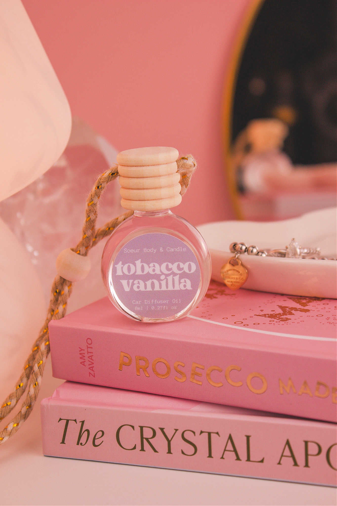 Tobacco Vanilla diffuser sitting on top of a stack of books with a jewelry dish in the background, set against a lamp, mirror, and crystals; fragrance notes: vanilla, amber, tobacco leaves, and tonka bean.