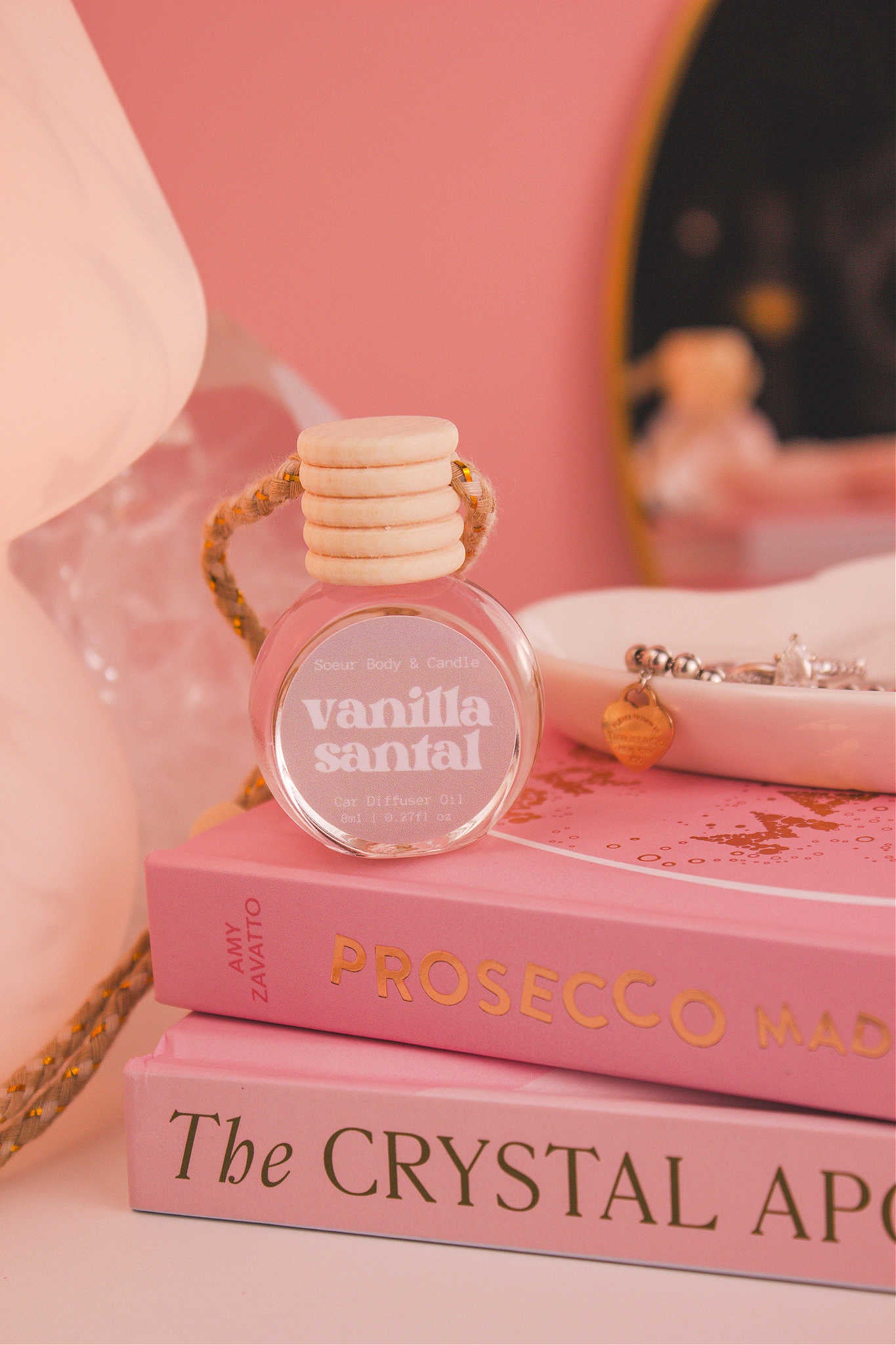 Vanilla Santal diffuser sitting on top of a stack of books with a jewelry dish in the background, set against a lamp, mirror, and crystals; fragrance notes: shea butter, coconut, vanilla, and sandalwood.