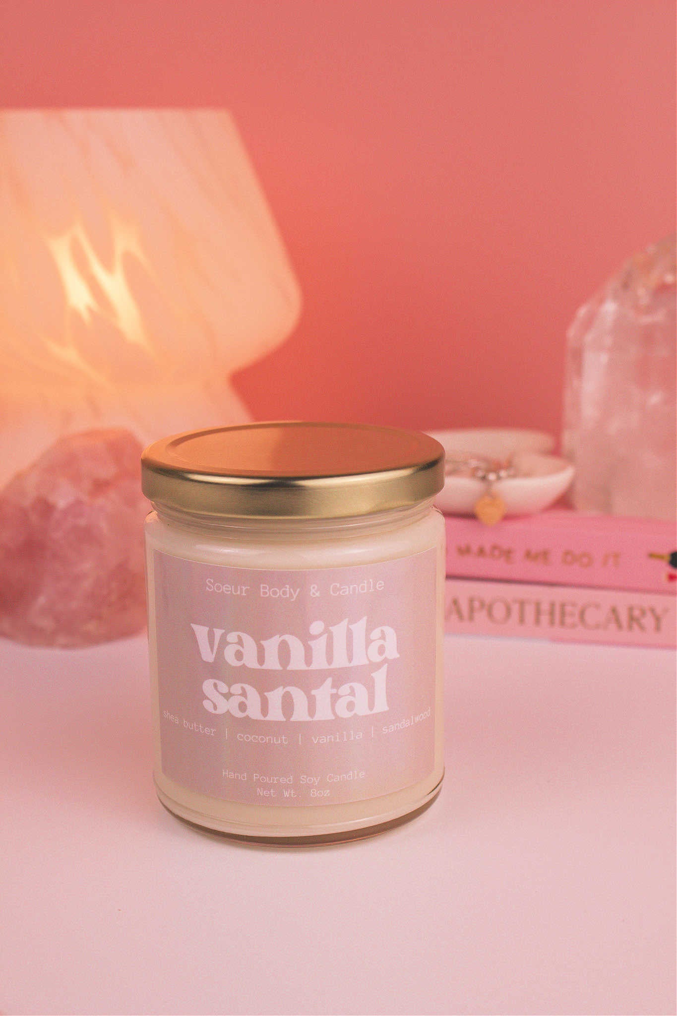Vanilla Santal candle with the lid on, with a stack of books and a jewelry dish in the background, set against a lamp and crystals; fragrance notes: shea butter, coconut, vanilla, and sandalwood.