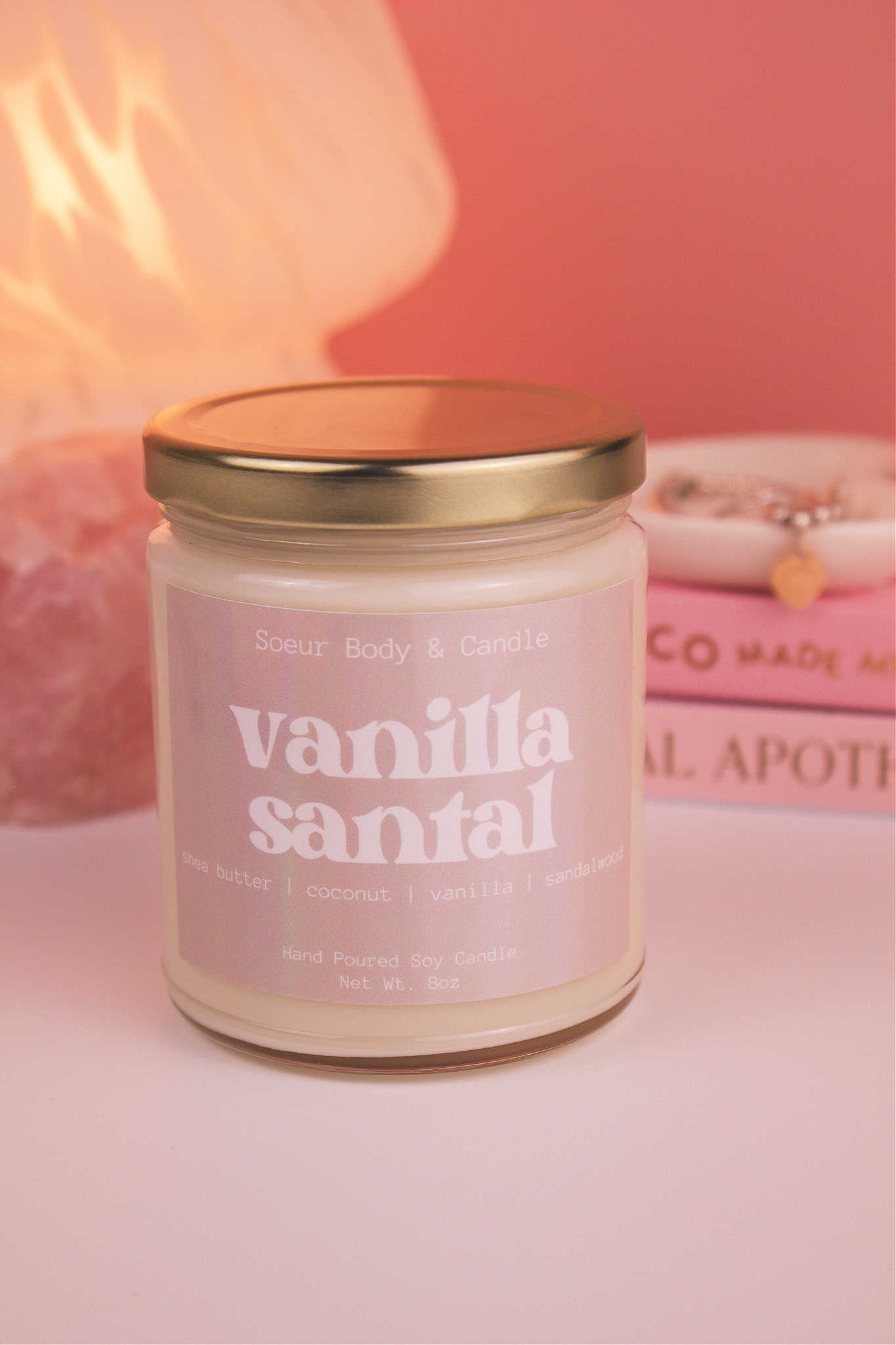 Close-up of Vanilla Santal candle with the lid on, with a stack of books and a jewelry dish in the background, set against a lamp and crystals; fragrance notes: shea butter, coconut, vanilla, and sandalwood.