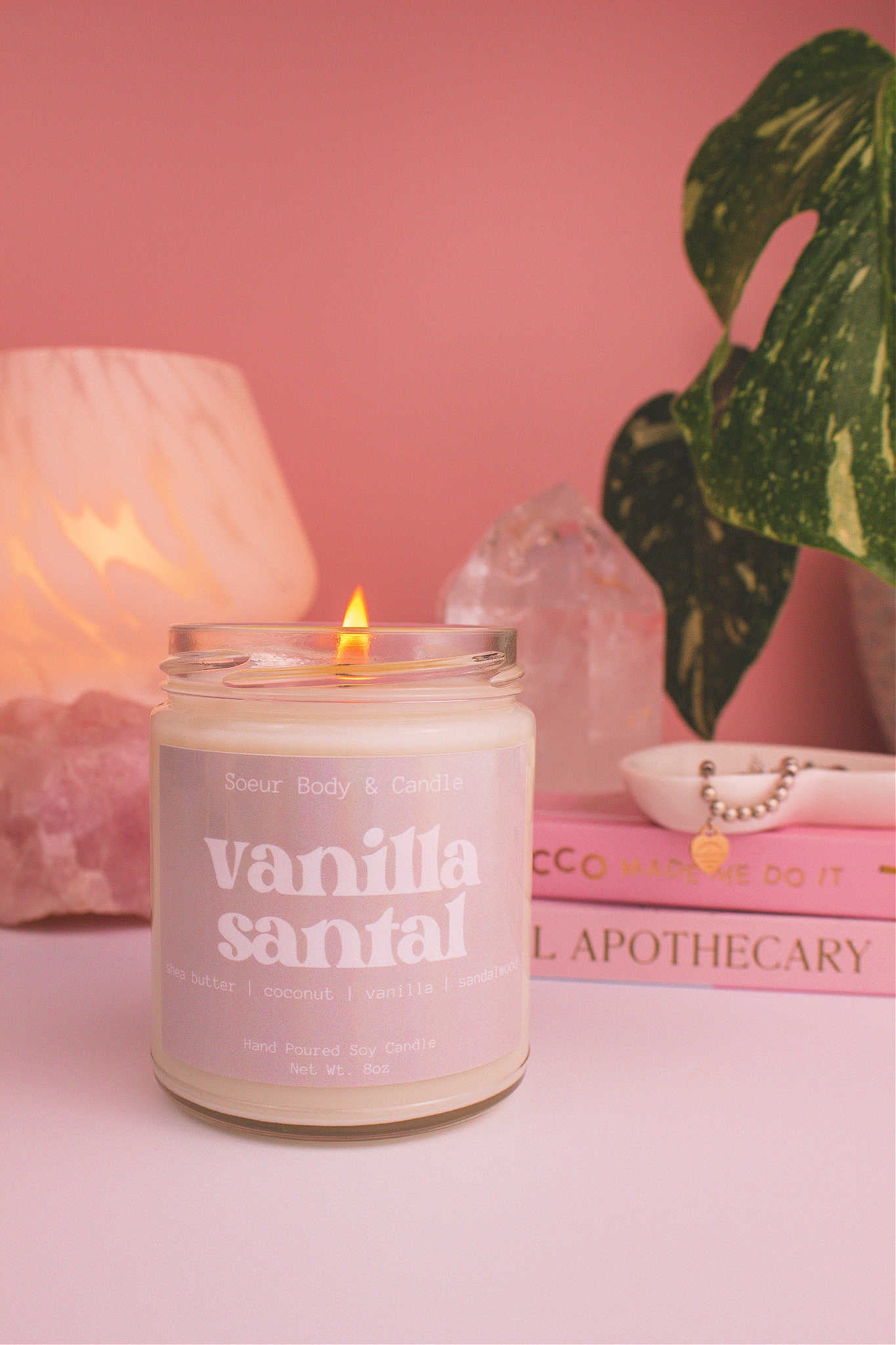 Vanilla Santal candle lit, with a stack of books and a jewelry dish in the background, set against a lamp, plant, and crystals; fragrance notes: shea butter, coconut, vanilla, and sandalwood.