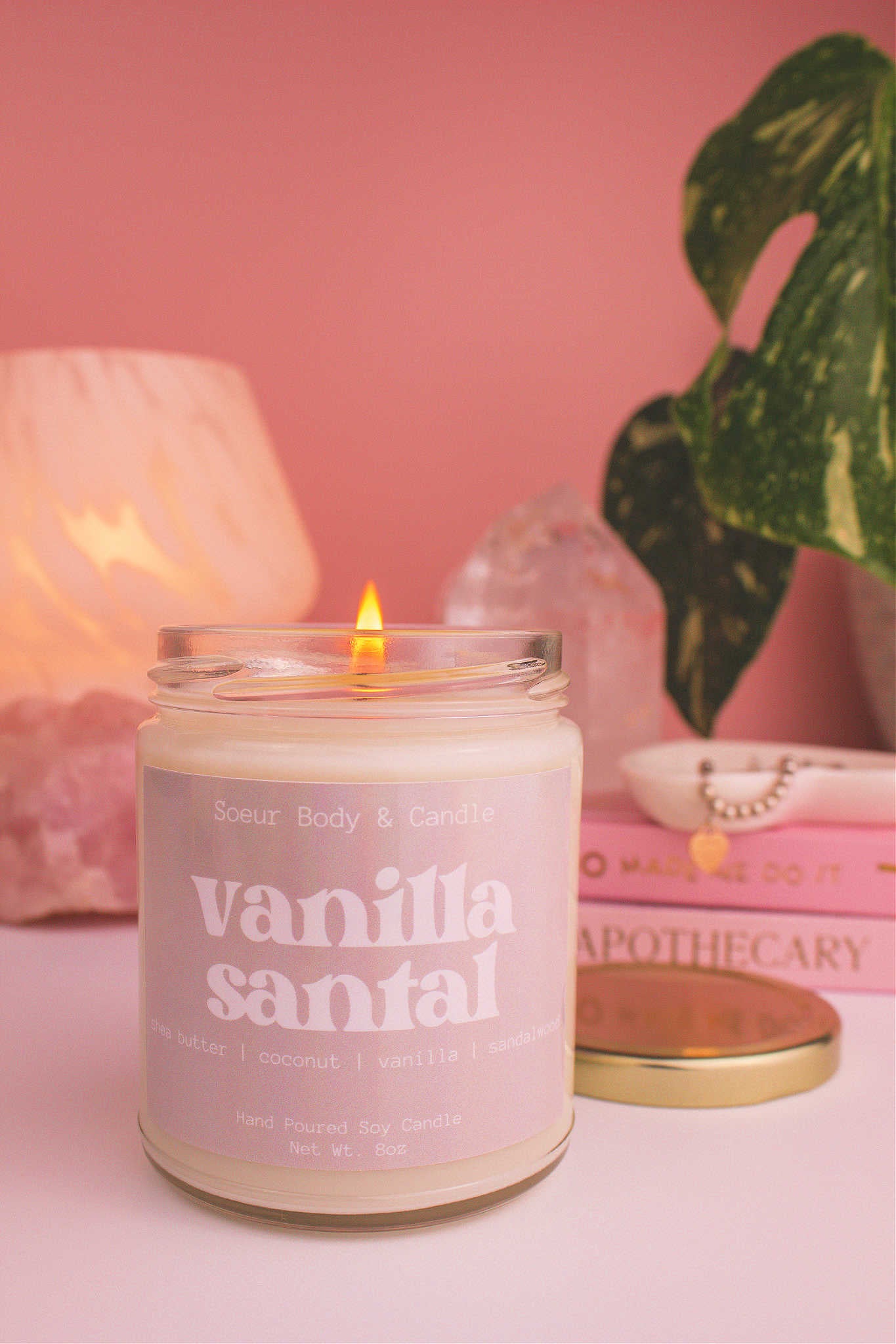Close-up of Vanilla Santal candle lit, with a stack of books and a jewelry dish in the background, set against a lamp, plant, and crystals; fragrance notes: shea butter, coconut, vanilla, and sandalwood.