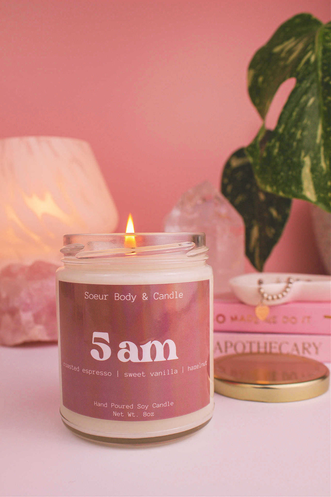 Close-up of 5am candle lit, with a stack of books and a jewelry dish in the background, set against a lamp, plant,  and crystals; fragrance notes: roasted espresso, sweet vanilla, hazelnut.