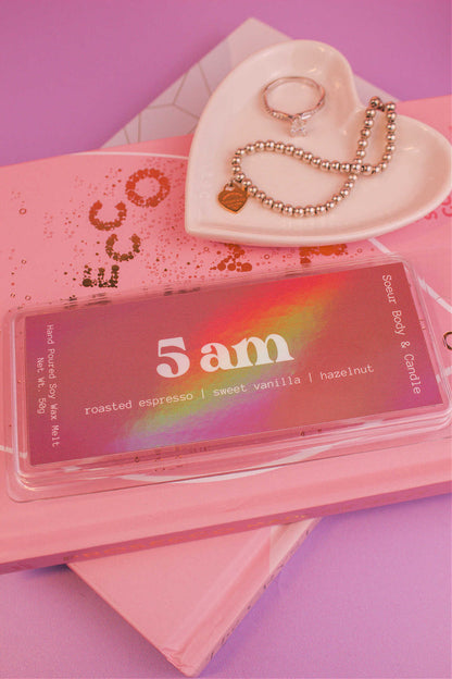 Close-up of the 5am wax melt on a stack of books with a jewelry dish in the background holding jewelry; fragrance notes: roasted espresso, sweet vanilla, and hazelnut.