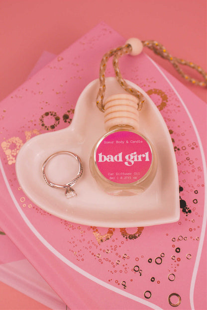 Bad Girl car diffuser in a jewelry dish with a ring beside it, sitting on top of books, showcasing bold notes of almond, coffee, jasmine, tonka bean, and cocoa.