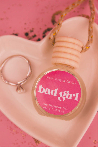 Close-up of Bad Girl car diffuser in a jewelry dish with a ring beside it, showcasing bold notes of almond, coffee, jasmine, tonka bean, and cocoa.