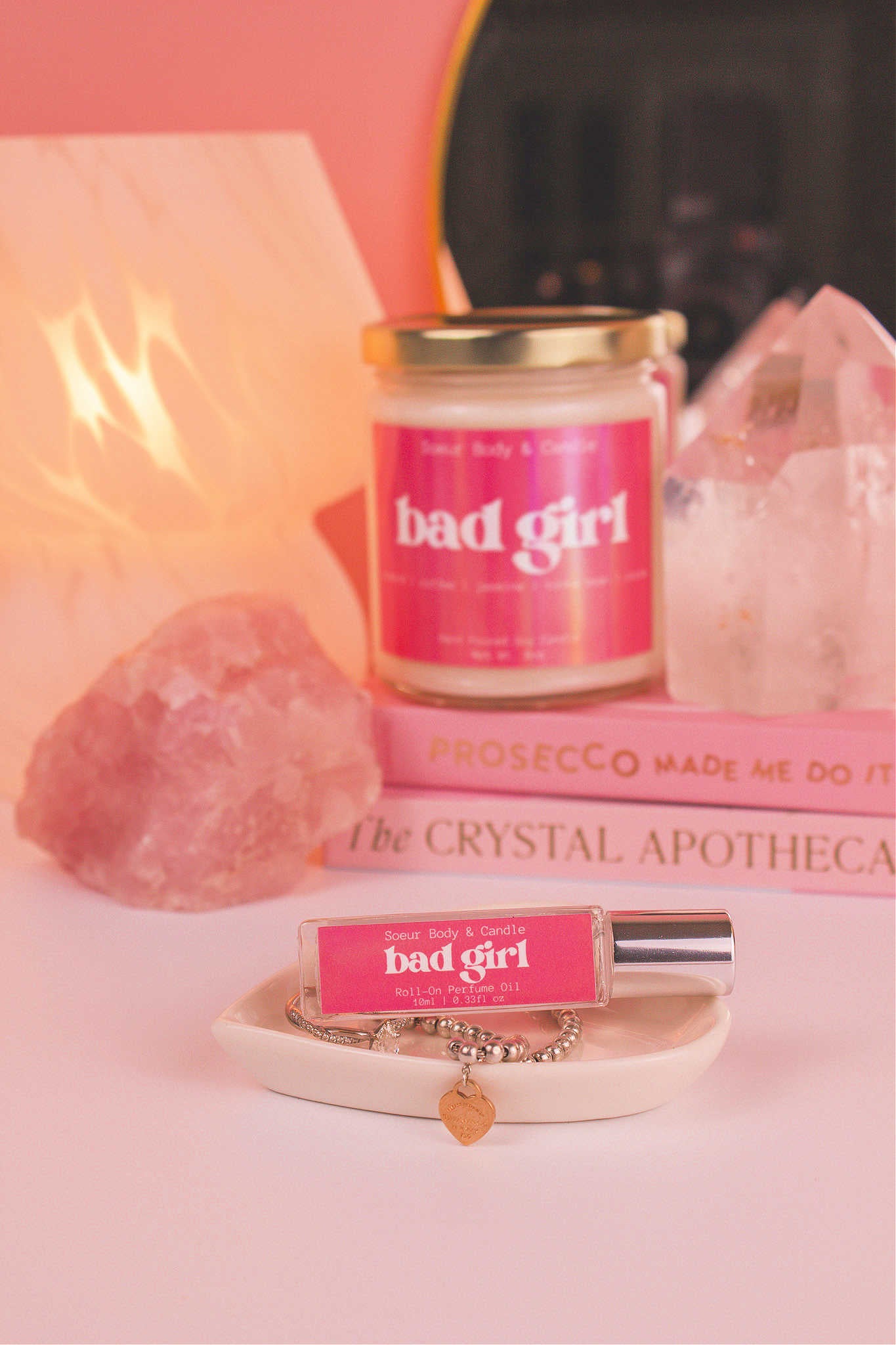 Bad Girl roll-on perfume elegantly displayed in a jewelry dish with coordinating jewelry. Matching candle, books, a lamp, and crystals in the background add a cozy vibe. Featuring notes of almond, coffee, jasmine, tonka bean, and cocoa.