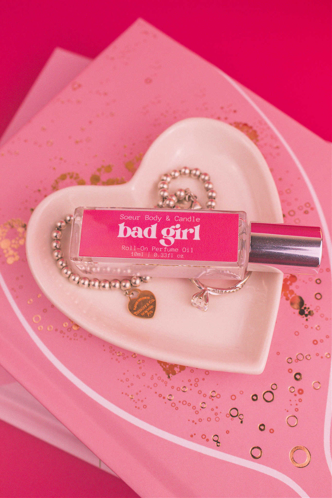 Bad Girl roll-on perfume bottle in a jewelry dish beside a ring and bracelet, highlighting its daring notes of almond, coffee, jasmine, tonka bean, and cocoa.