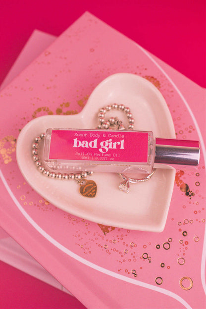 Bad Girl roll-on perfume bottle in a jewelry dish beside a ring and bracelet, highlighting its daring notes of almond, coffee, jasmine, tonka bean, and cocoa.