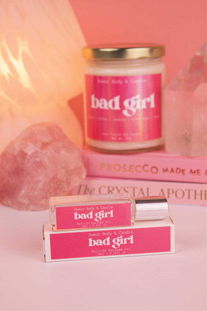 Focused shot of Bad Girl roll-on perfume in the foreground with matching candle softly blurred in the background, along with books, a lamp, and crystals, adding a cozy vibe.  Featuring notes of almond, coffee, jasmine, tonka bean, and cocoa.