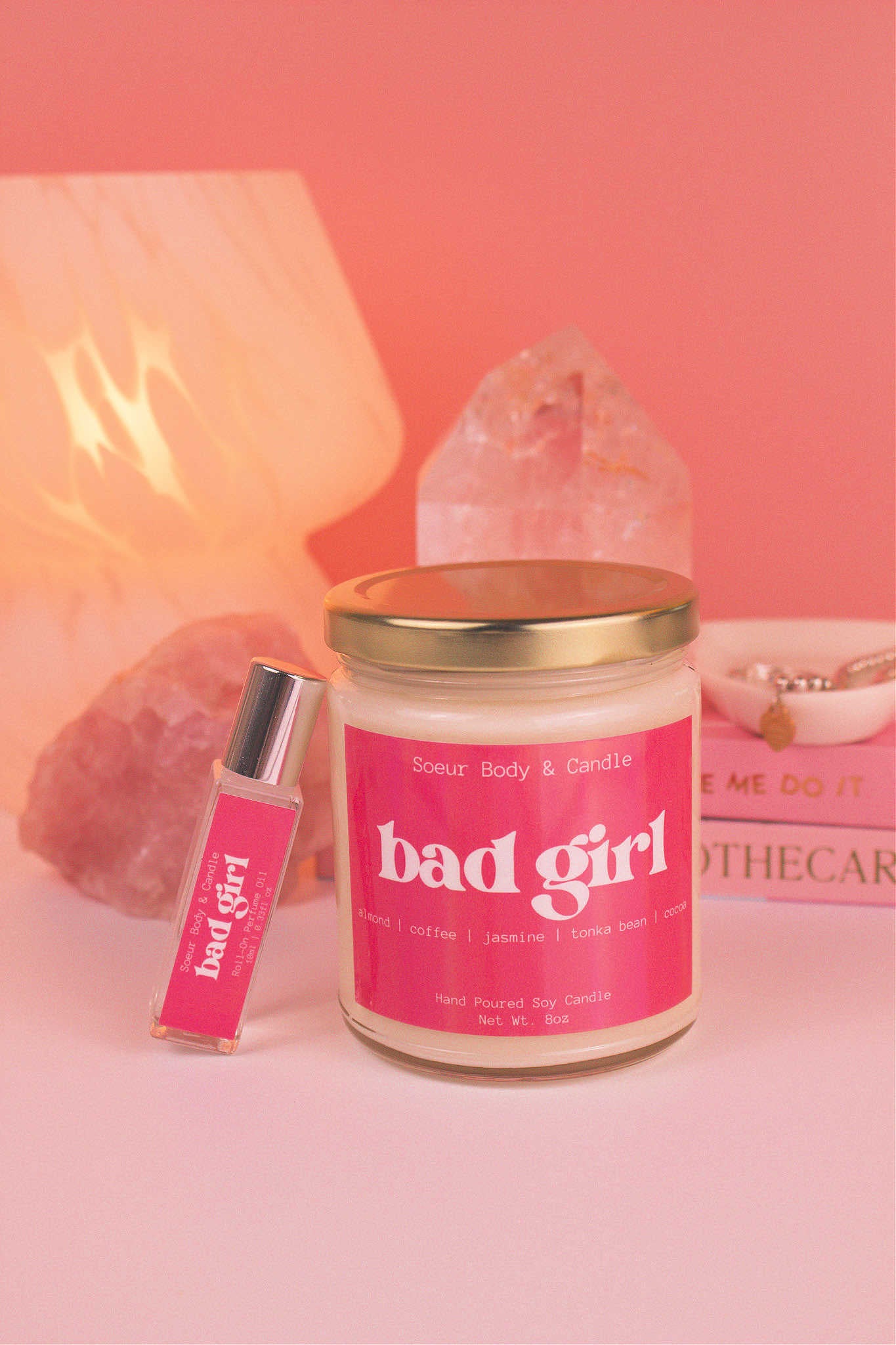 Bad Girl roll-on perfume and candle together, with books, a lamp, and crystals in the background, adding a cozy vibe. Featuring a bold fusion of almond, coffee, jasmine, tonka bean, and cocoa. 