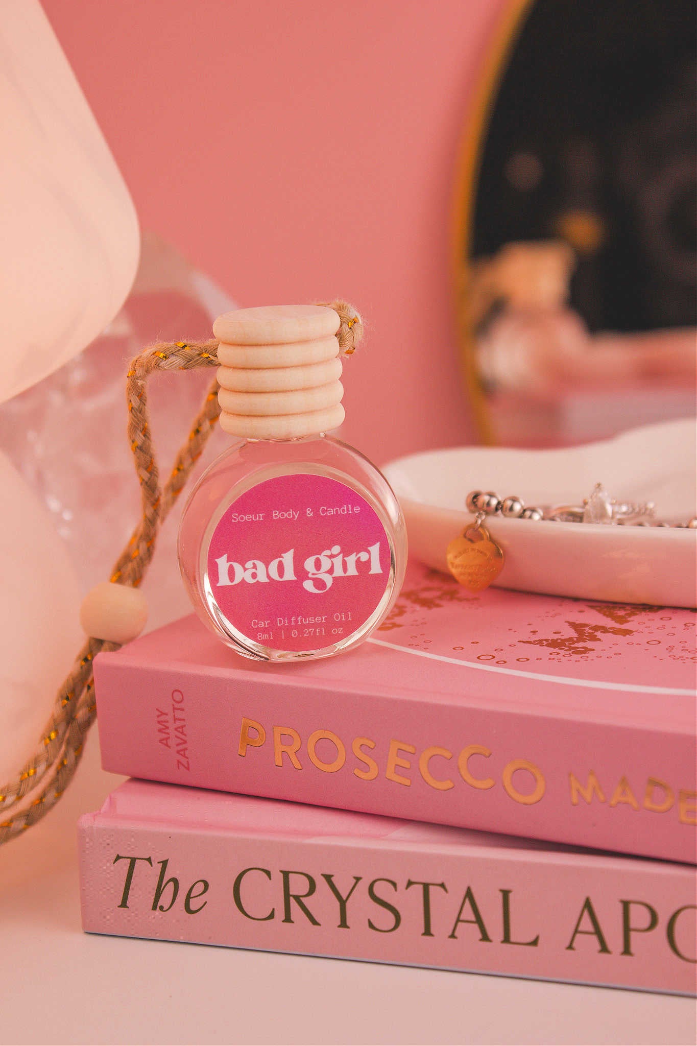 Bad Girl diffuser sitting on top of a stack of books with a jewelry dish in the background, set against a lamp, mirror, and crystals; fragrance notes:  almond, coffee, jasmine, tonka bean, and cocoa.