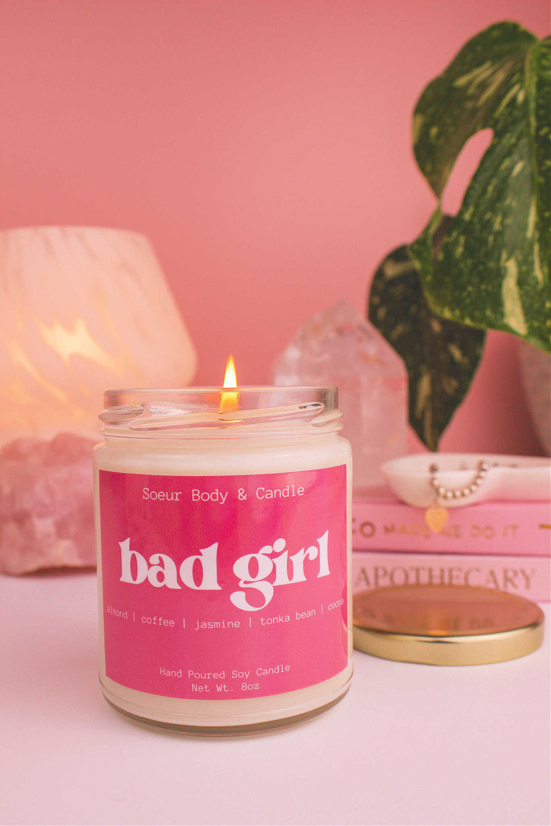 Close-up of Bad Girl candle lit, with a jewelry dish on top of books and a lamp, plant, and crystals in the background. Featuring notes of  almond, coffee, jasmine, tonka bean, and cocoa notes.