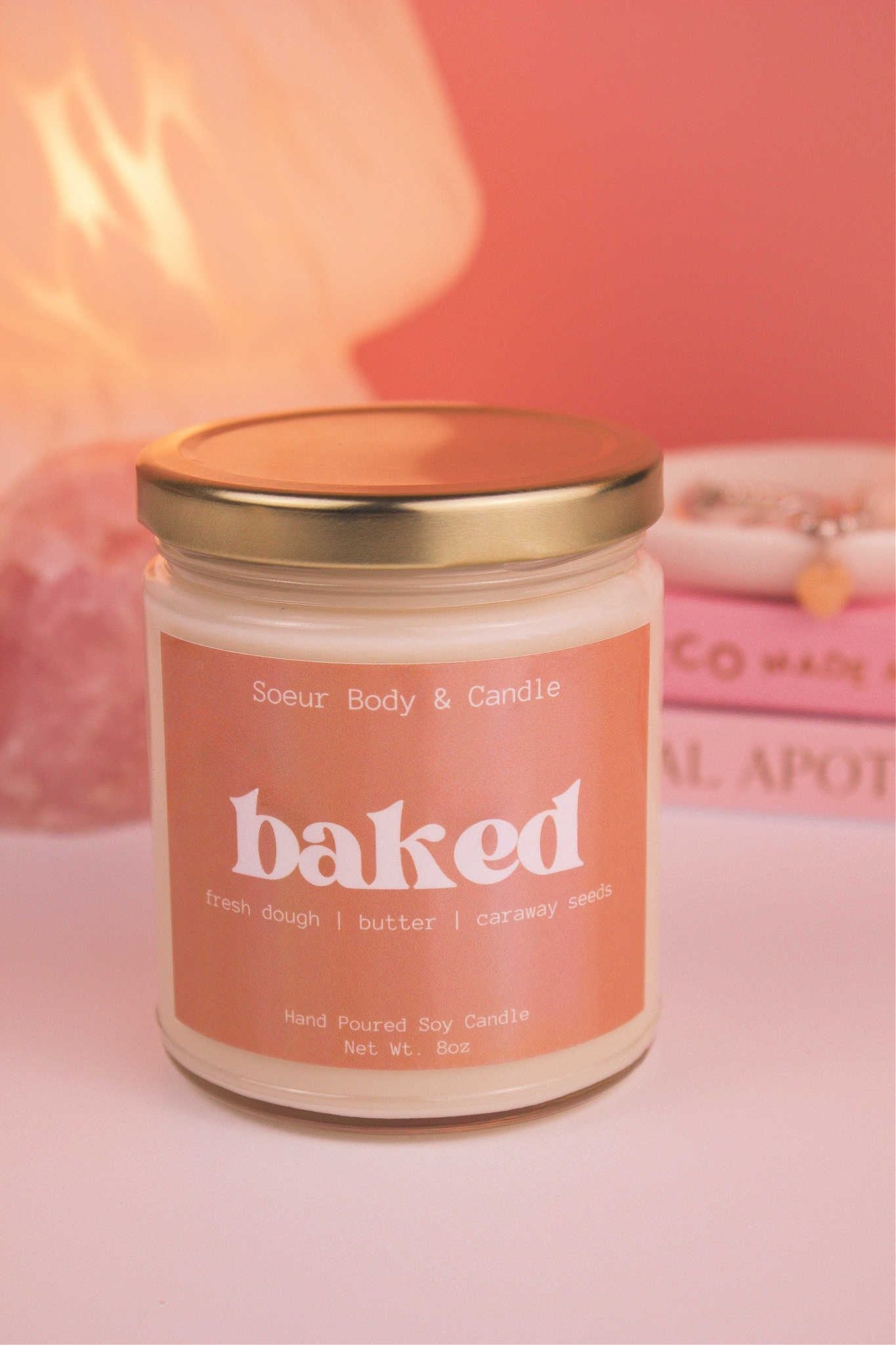 Close-up of Baked candle with the lid on, with a stack of books and a jewelry dish in the background, set against a lamp and crystals; fragrance notes: fresh dough, butter, and caraway seeds.