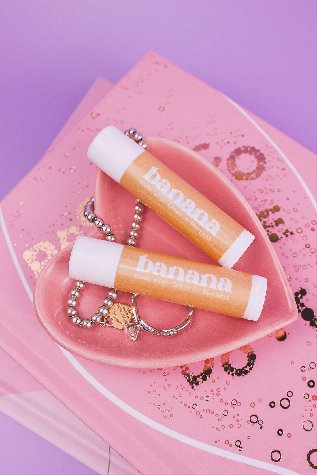 Two Banana flavoured lip balms displayed in a jewelry dish with a ring and bracelet, sitting on top of books.