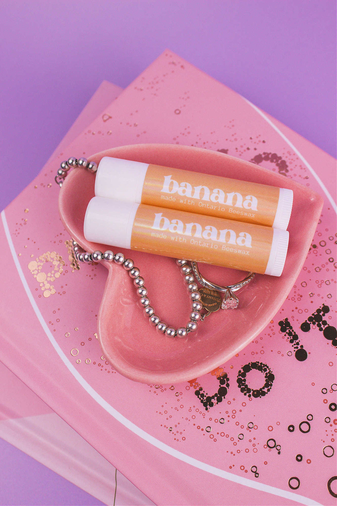 Two Banana flavoured lip balms displayed in a jewelry dish with a ring and bracelet, sitting on top of books.