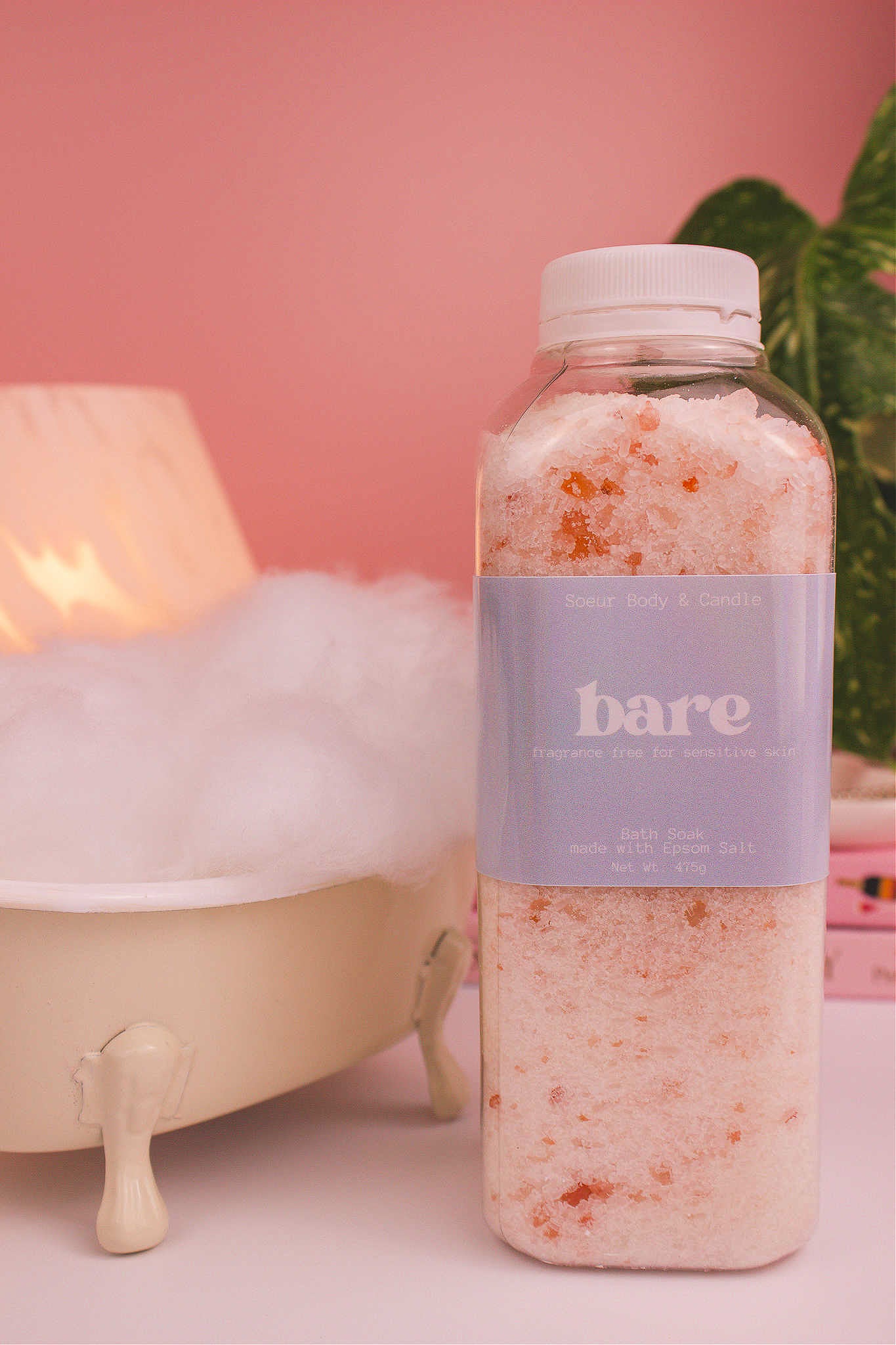 Bare bath soak beside a small tub with bubbles, a lamp, and plants in the background, creating a relaxing vibe. Fragrance free for sensitive skin.