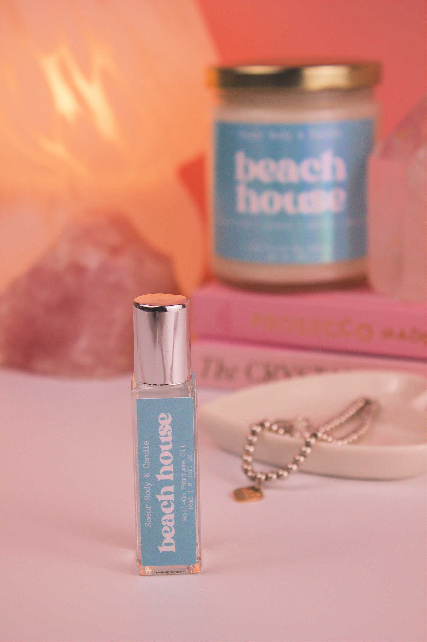 Focused shot of Beach House roll-on perfume in the foreground with its matching candle softly blurred in the background, along with books, a lamp, and crystals, adding a cozy vibe. Featuring notes of citrus fruits, coconut, vanilla, and warm musk.