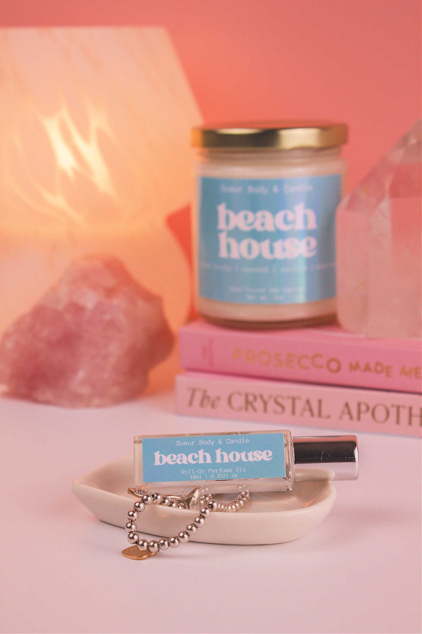 Beach House roll-on perfume elegantly set in a jewelry dish with jewelry, and matching candle, books, lamp, and crystals blurred in the background, adding a cozy vibe. Featuring notes of citrus fruits, coconut, vanilla, and warm musk.