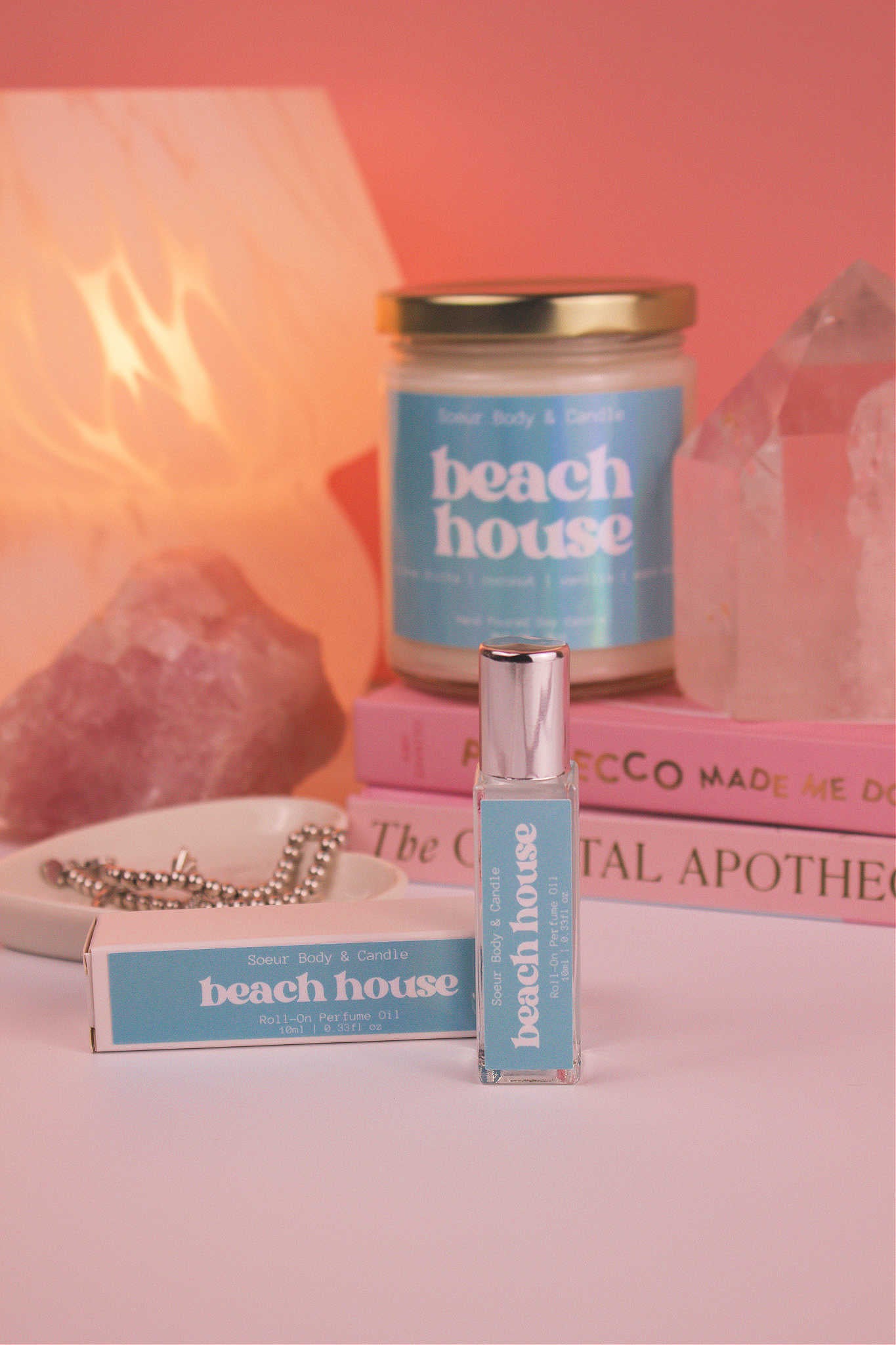 Focused shot of Beach House roll-on perfume and its packaging in the foreground, with its matching candle and jewelry dish softly blurred in the background, along with books, a lamp, and crystals, adding a cozy vibe. Featuring notes of citrus fruits, coconut, vanilla, and warm musk.