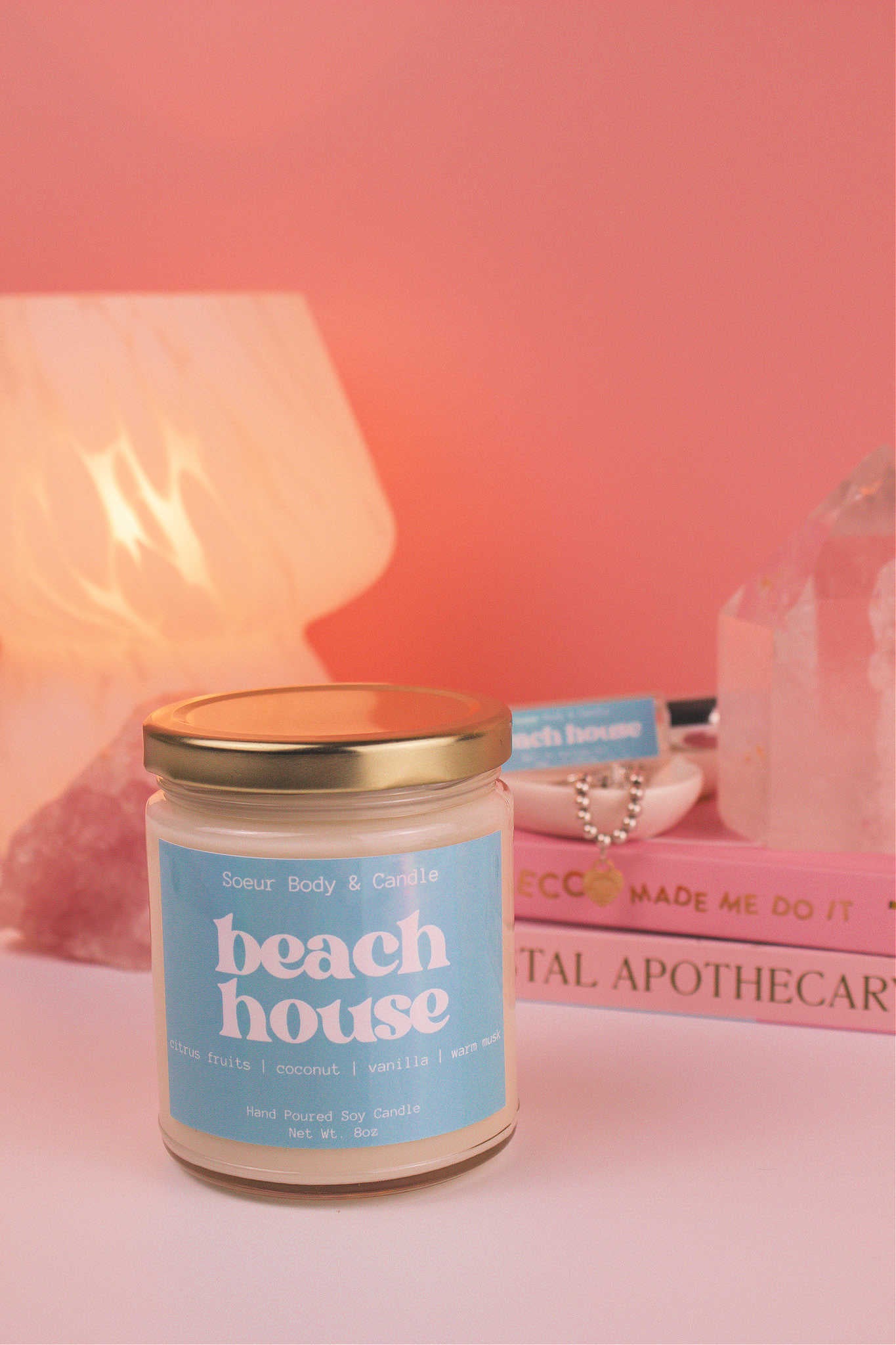 Beach House candle with the lid on, with a stack of books and a jewelry dish in the background, set against a lamp and crystals; fragrance notes: citrus fruits, coconut, vanilla, and warm musk.