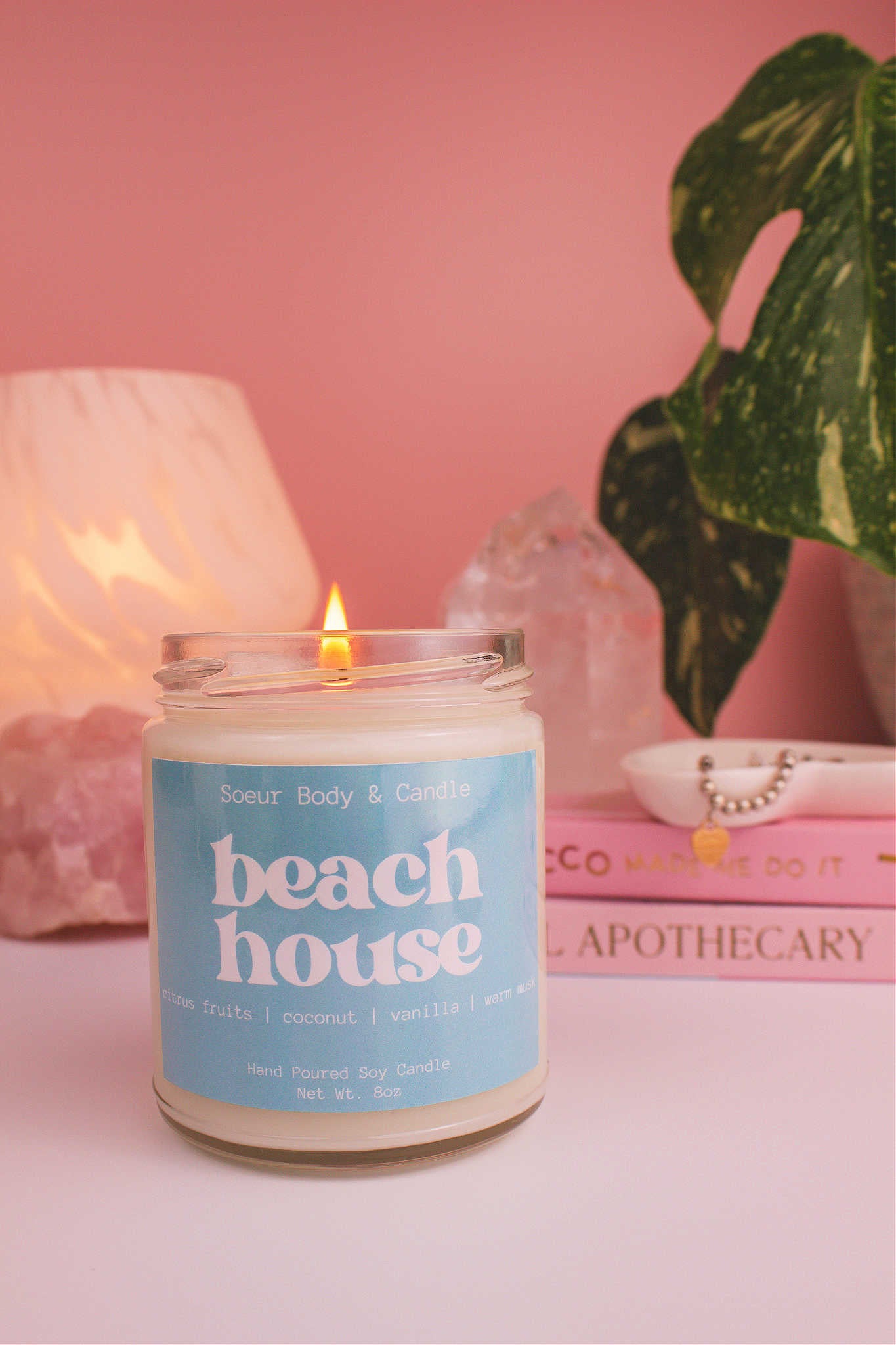 Beach House candle lit, with a jewelry dish, books, lamp, plant, and crystals in the background. Featuring notes of citrus fruits, coconut, vanilla, and warm musk.