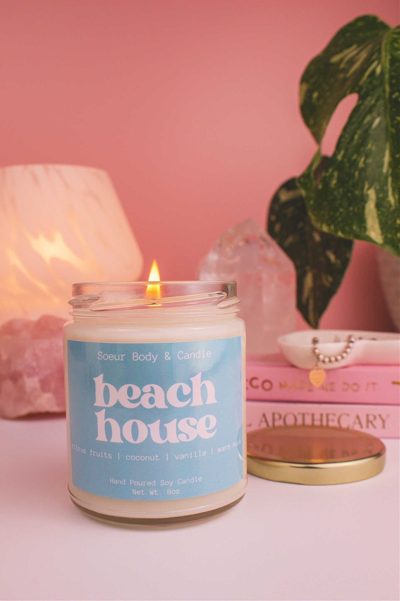 Close-up of Beach House candle lit, with a jewelry dish, books, lamp, plant, and crystals in the background. Featuring refreshing notes of citrus fruits, coconut, vanilla, and warm musk.