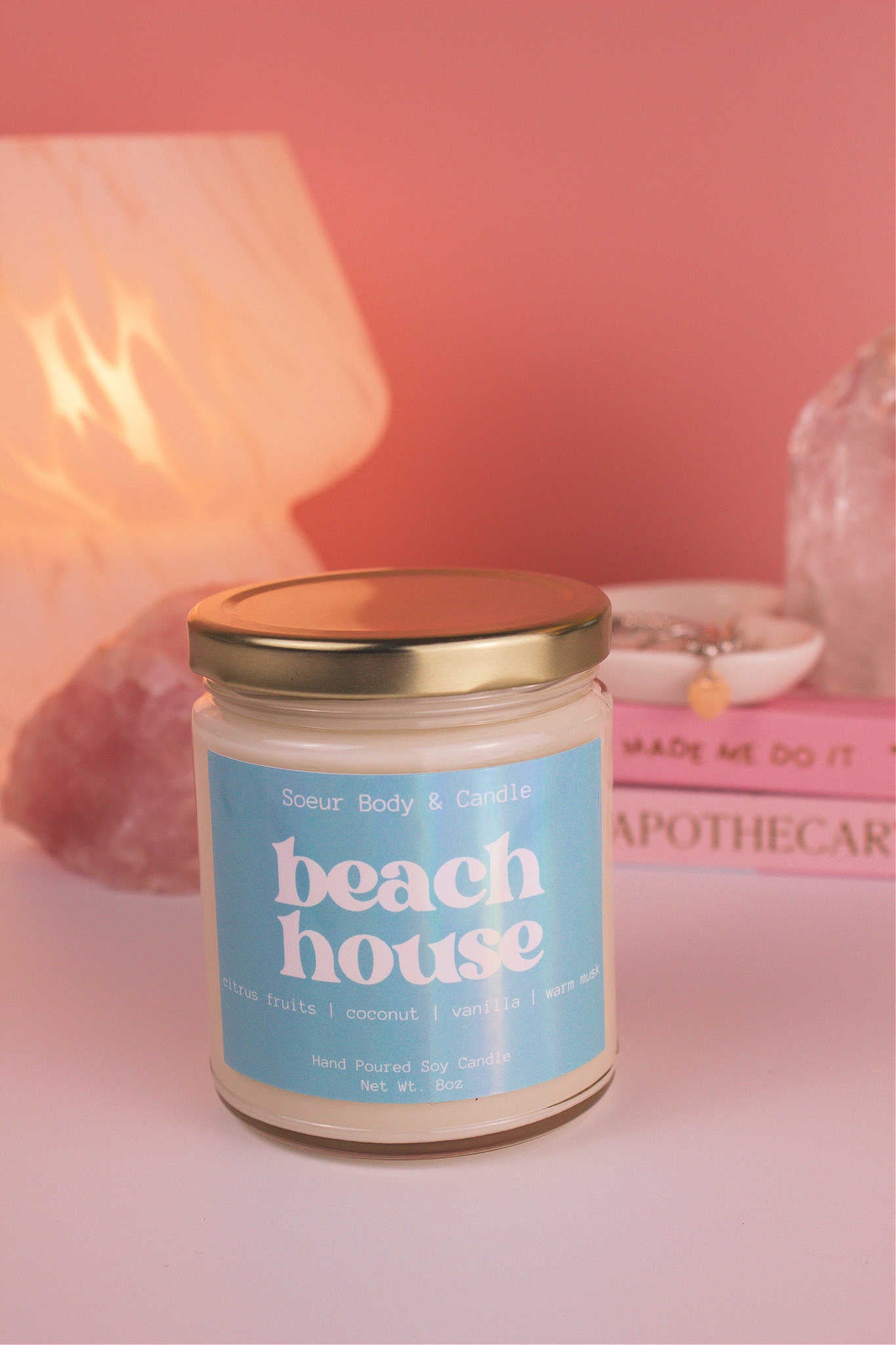 Close-up of Beach House candle with the lid on, with a stack of books and a jewelry dish in the background, set against a lamp and crystals; fragrance notes: citrus fruits, coconut, vanilla, and warm musk.