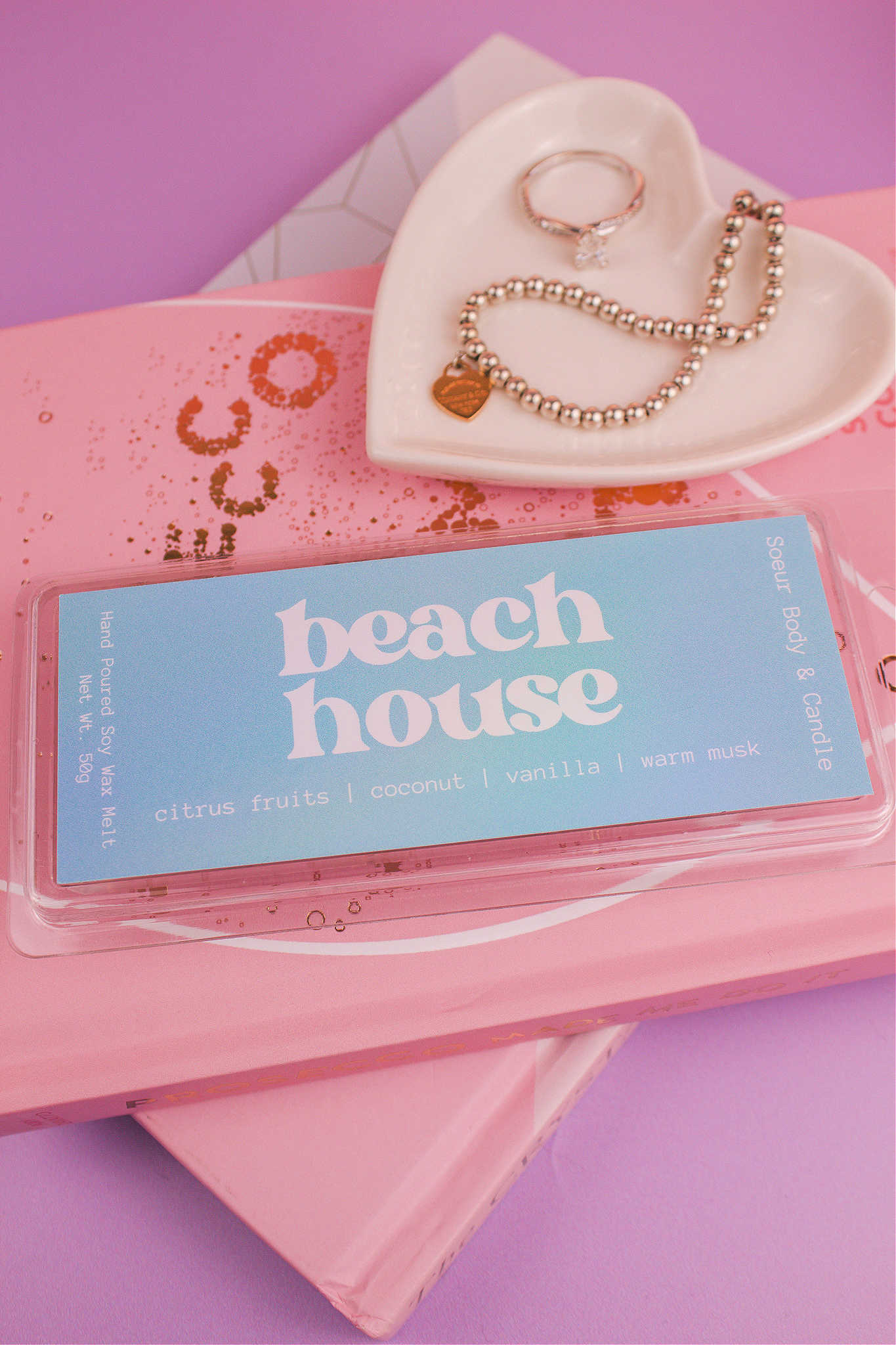 Beach House wax melt on a stack of books with a jewelry dish in the background holding jewelry; fragrance notes: citrus fruits, coconut, vanilla, and warm musk.