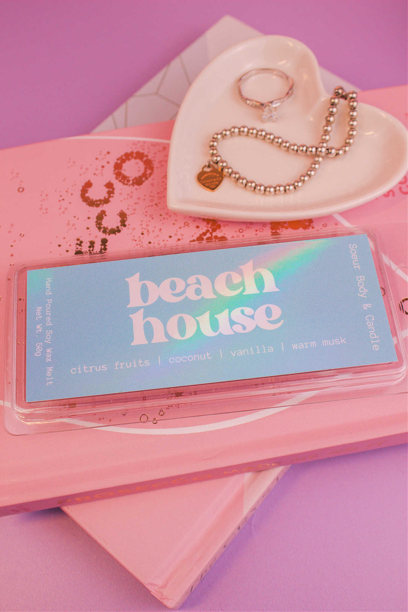 Close-up of the Beach House wax melt on a stack of books with a jewelry dish in the background holding jewelry; fragrance notes: citrus fruits, coconut, vanilla, and warm musk.