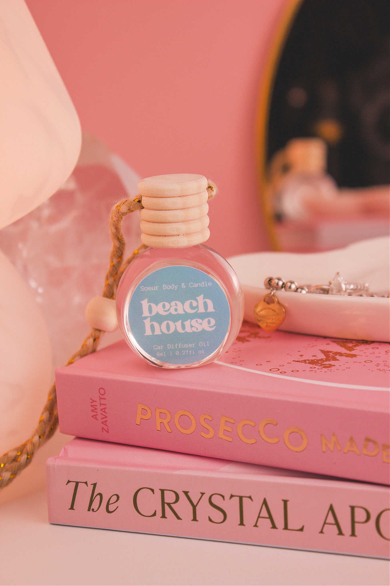 Beach House diffuser sitting on top of a stack of books with a jewelry dish in the background, set against a lamp, mirror, and crystals; fragrance notes:  citrus fruits, coconut, vanilla, and warm musk.