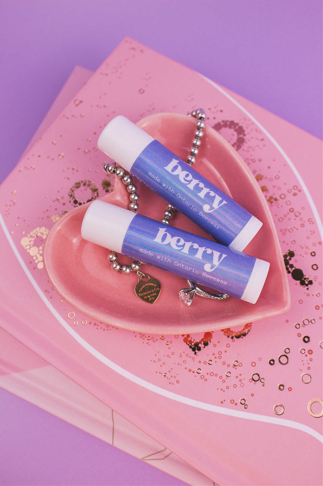 Two Berry flavoured lip balms displayed in a jewelry dish with a ring and bracelet, sitting on top of books.