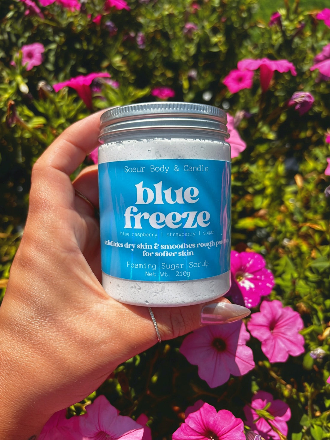 A hand holding a jar of Soeur Body and Candle Blue Freeze Foaming Sugar Scrub against a background of green foliage and pink flowers.