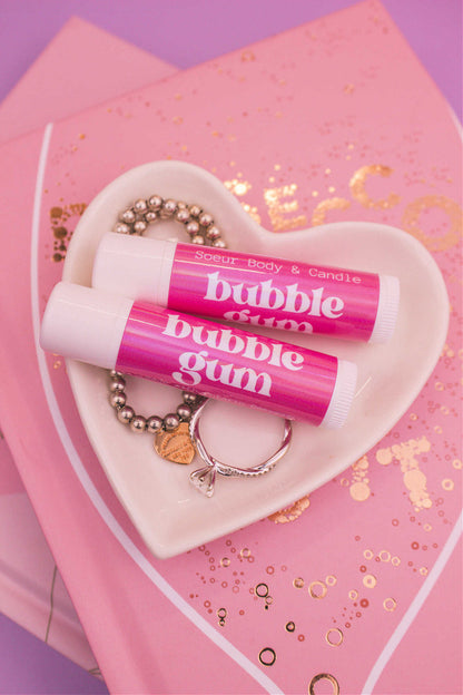 Two Bubblegum flavoured lip balms displayed in a jewelry dish with a ring and bracelet, sitting on top of books.