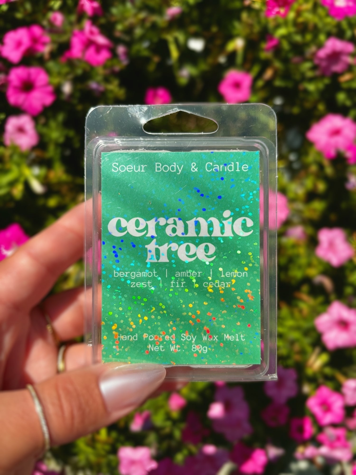 A hand holding a "Ceramic Tree" Soy Wax Melt with pink flowers and green foliage in the background. 