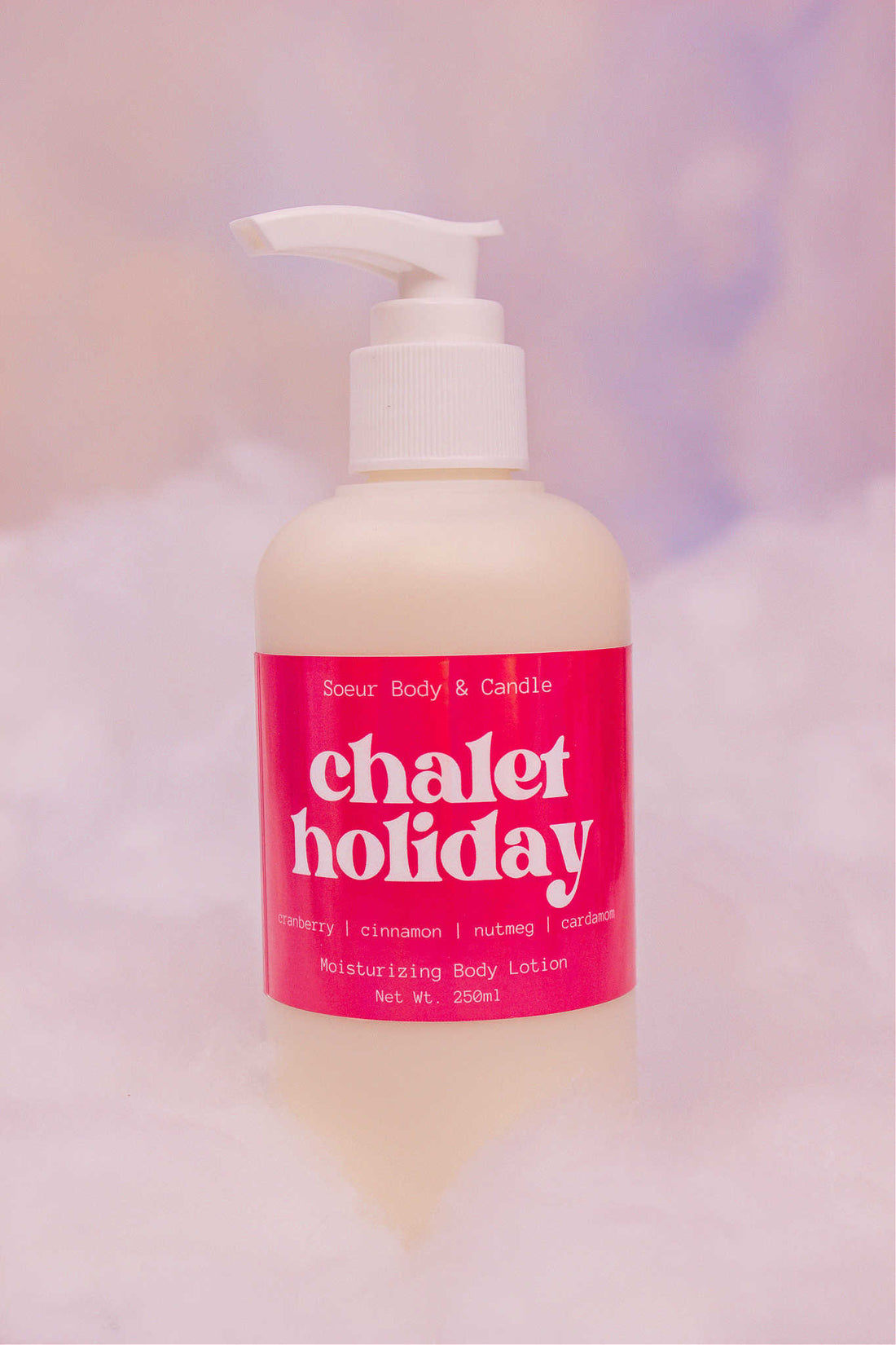 Chalet Holiday body lotion sitting in fake snow against a cloudy background. Featuring notes of cranberry, cinnamon, nutmeg, and cardamom.