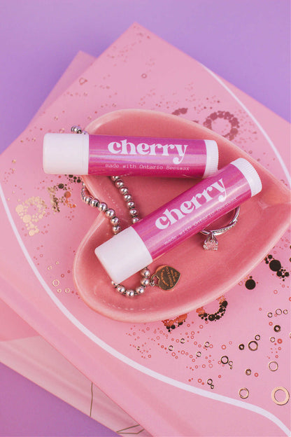 Two Cherry flavoured lip balms displayed in a jewelry dish with a ring and bracelet, sitting on top of books.