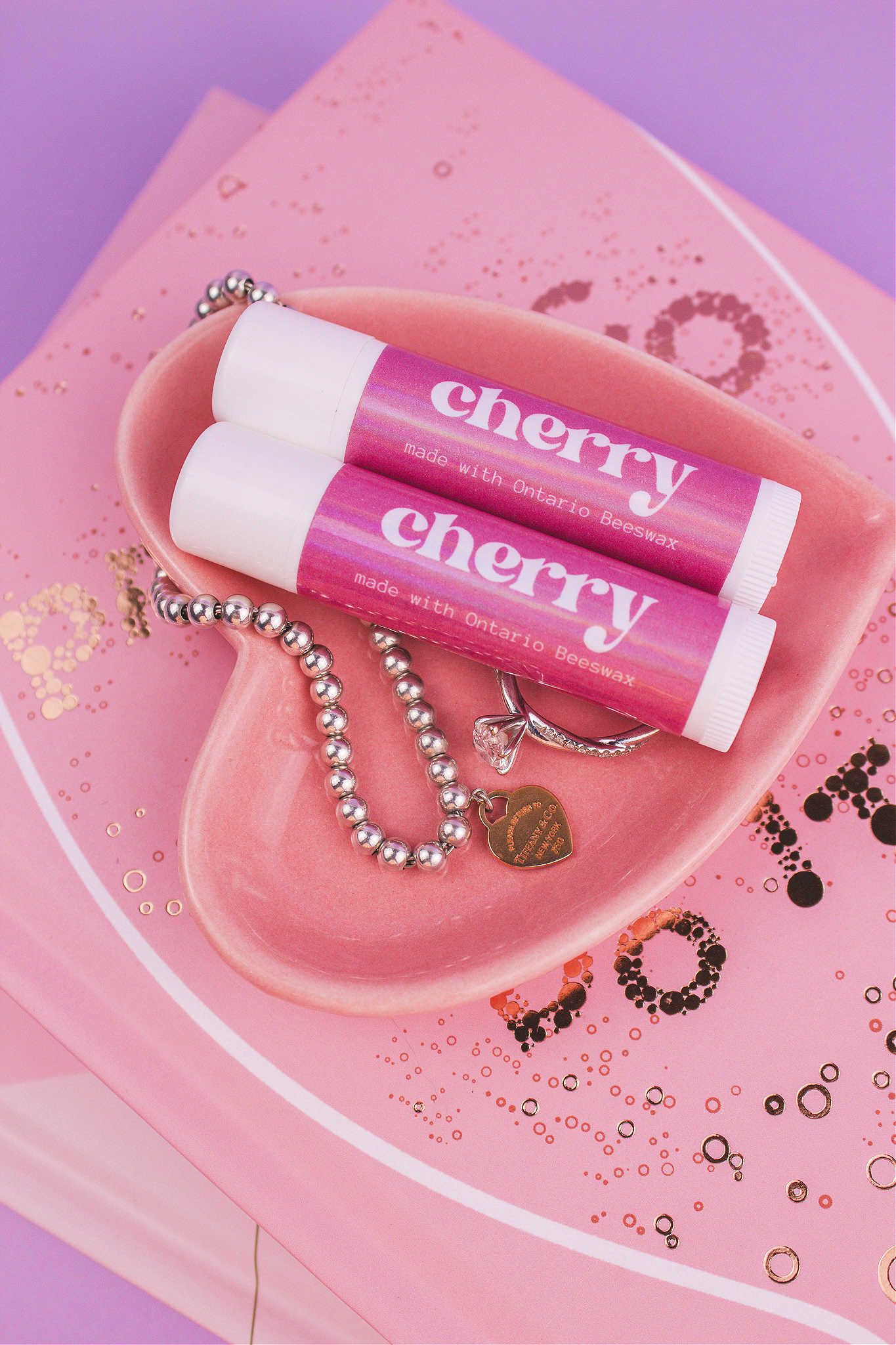 Two Cherry flavoured lip balms displayed in a jewelry dish with a ring and bracelet, sitting on top of books.