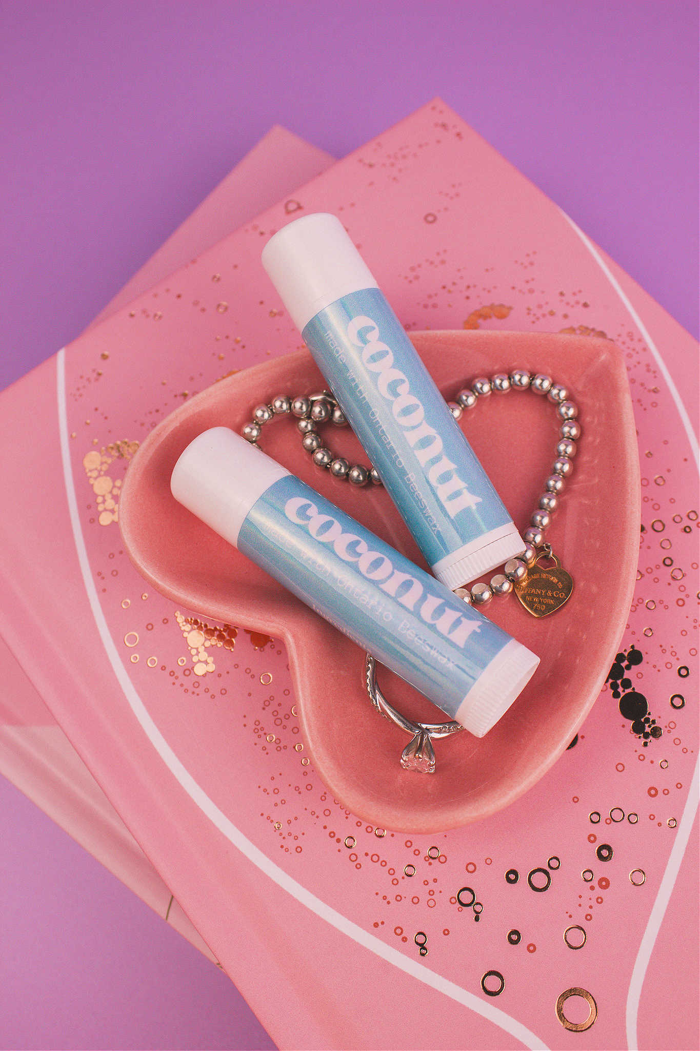 Two Coconut flavoured lip balms displayed in a jewelry dish with a ring and bracelet, sitting on top of books.