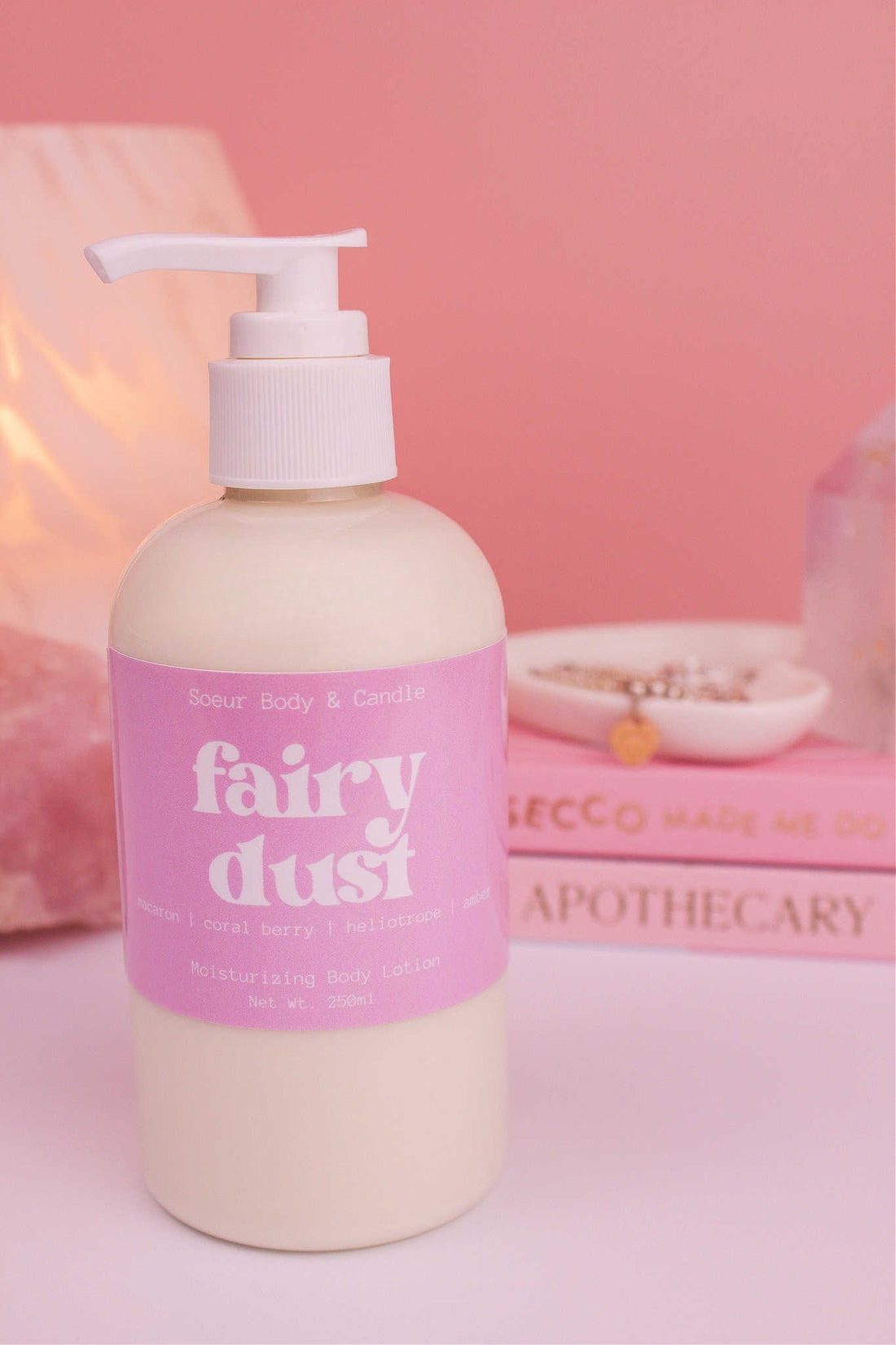 Close-up of Fairy Dust body lotion, with a stack of books, a jewelry dish, lamp, and crystal in the background; fragrance notes: macaron, coral berry, heliotrope, and amber.