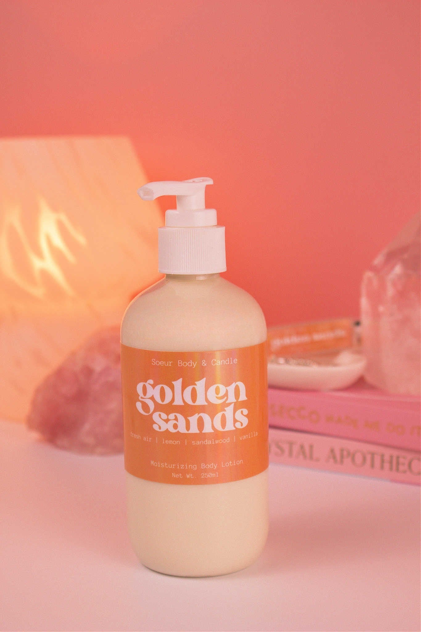 Golden Sands body lotion with a stack of books, a jewelry dish, lamp, and crystals in the background; fragrance notes: fresh air, lemon, sandalwood, and vanilla.